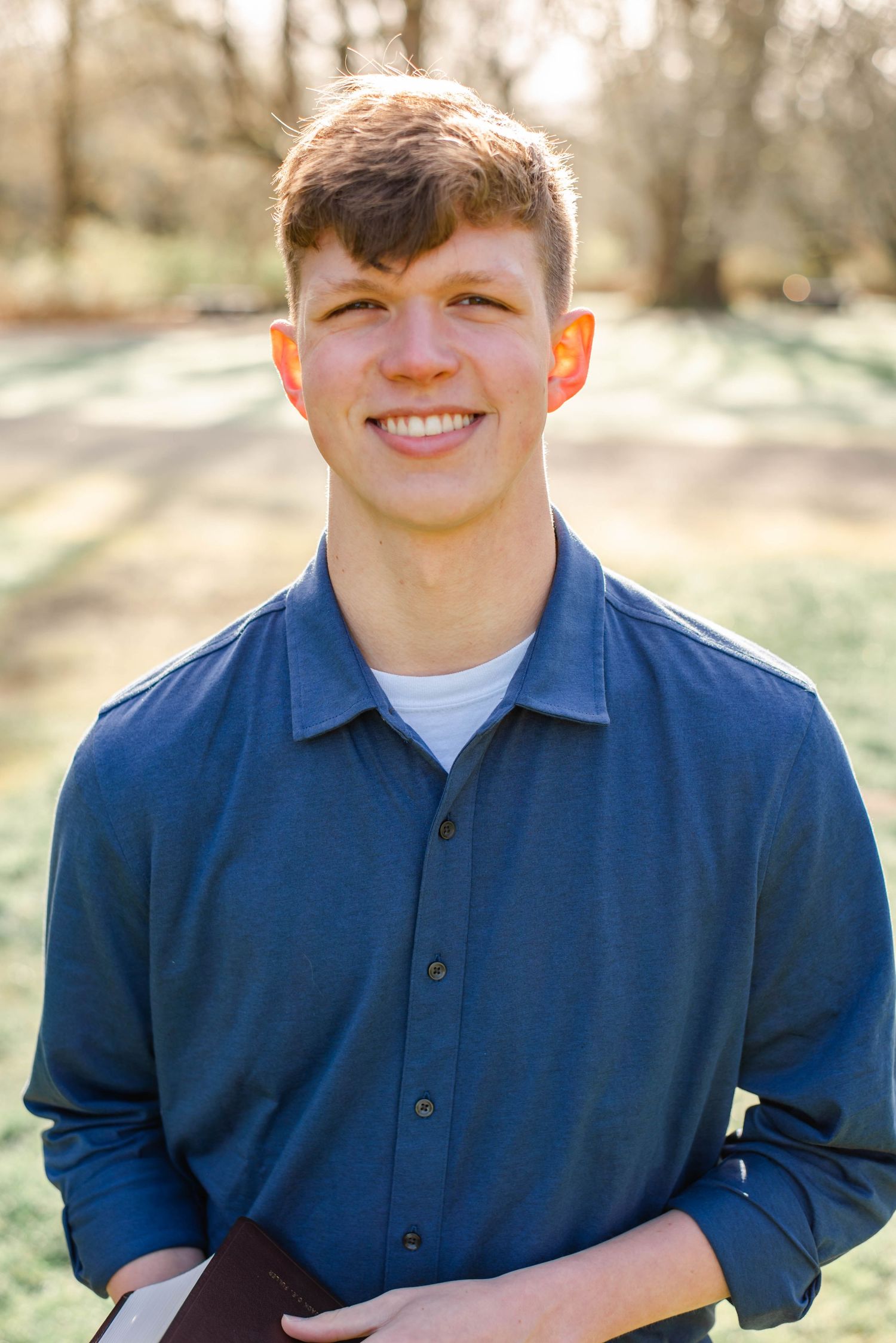 Smiling young man in blue button-up shirt holding a book outdoors in a sunny park setting.