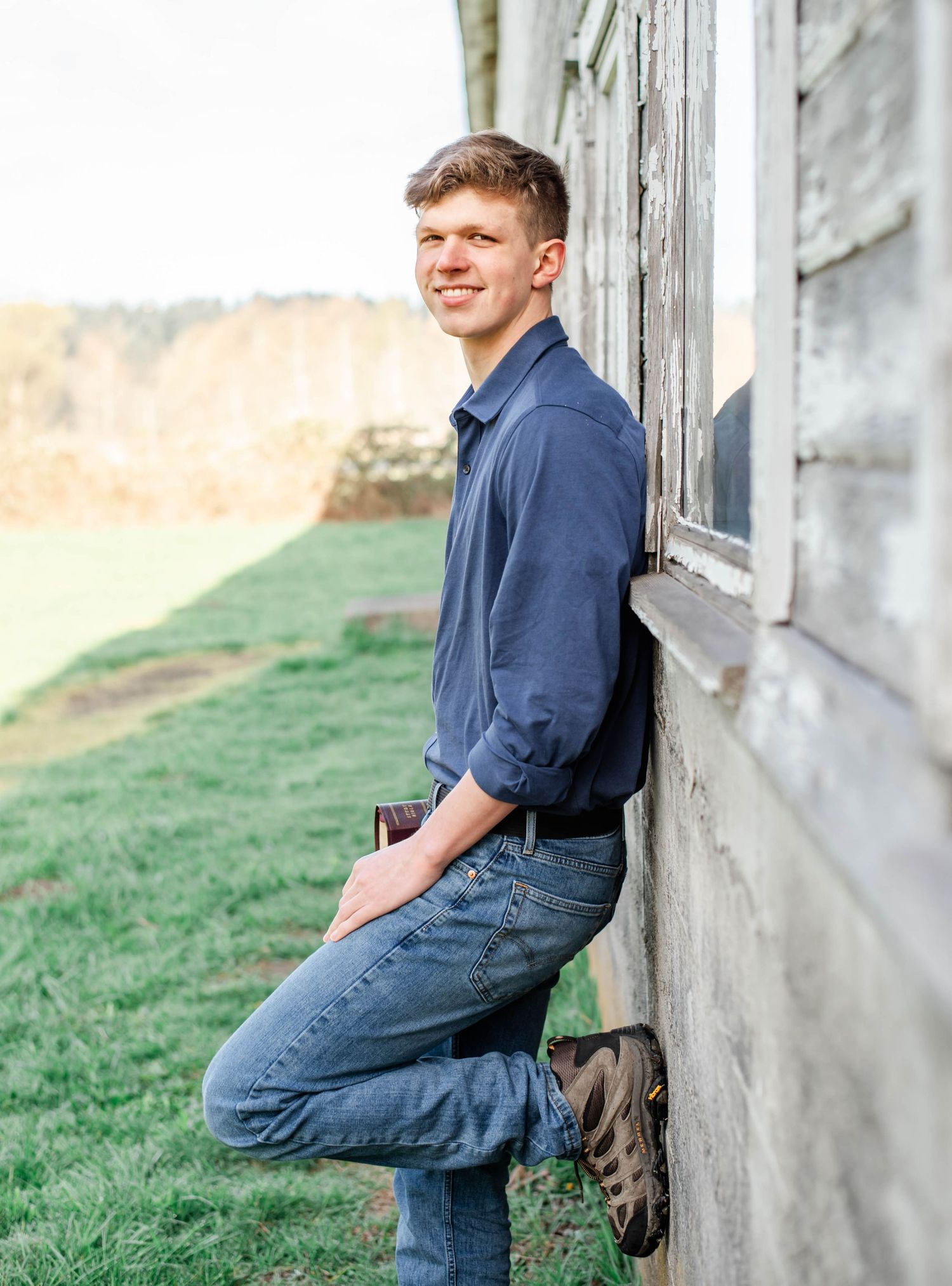 Young man in blue shirt and jeans leaning against rustic barn wall, smiling outdoors on green farm.