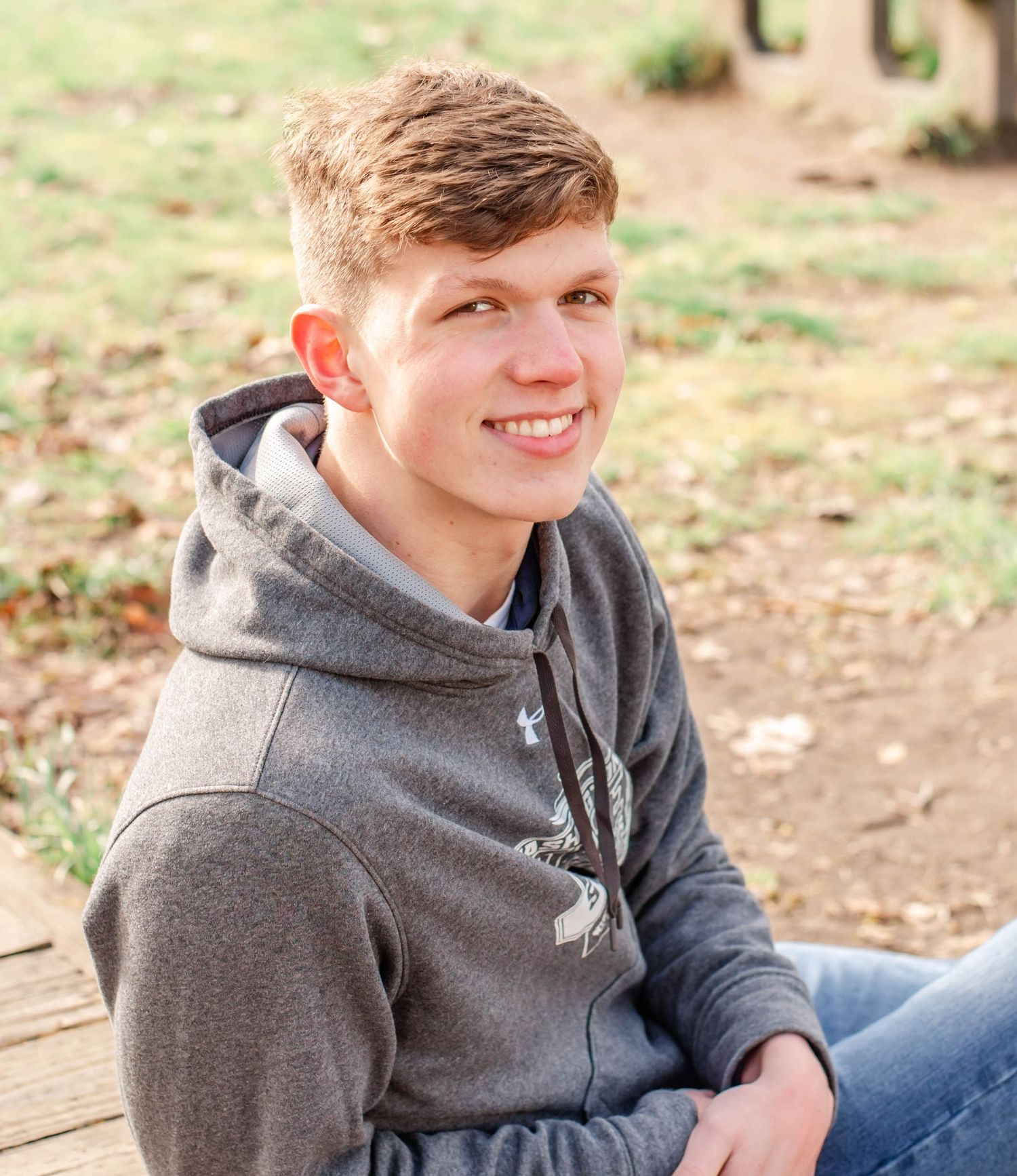 Smiling teenage boy in gray hoodie sitting outdoors on wooden surface with trees in background.