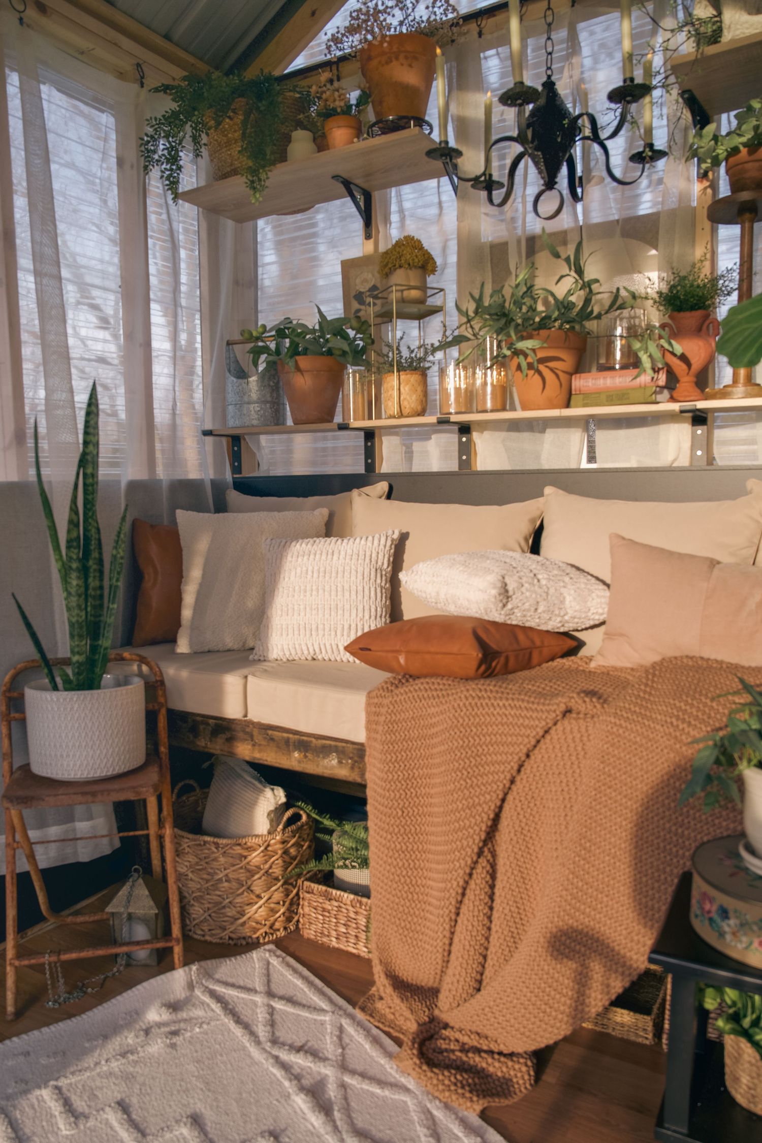 Cozy boho greenhouse nook with pallet sofa, earth-tone pillows, knit throw, and lush potted plants on shelves.