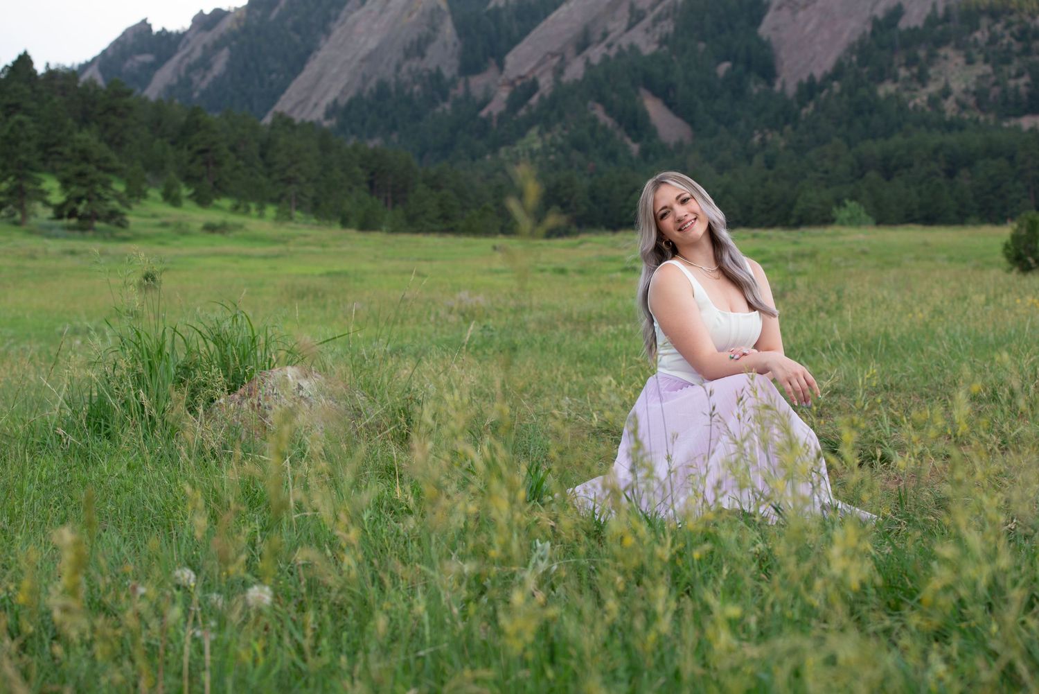 A high school senior woman in a white dress sits in a lush green meadow with the Flatirons mountains in the background.