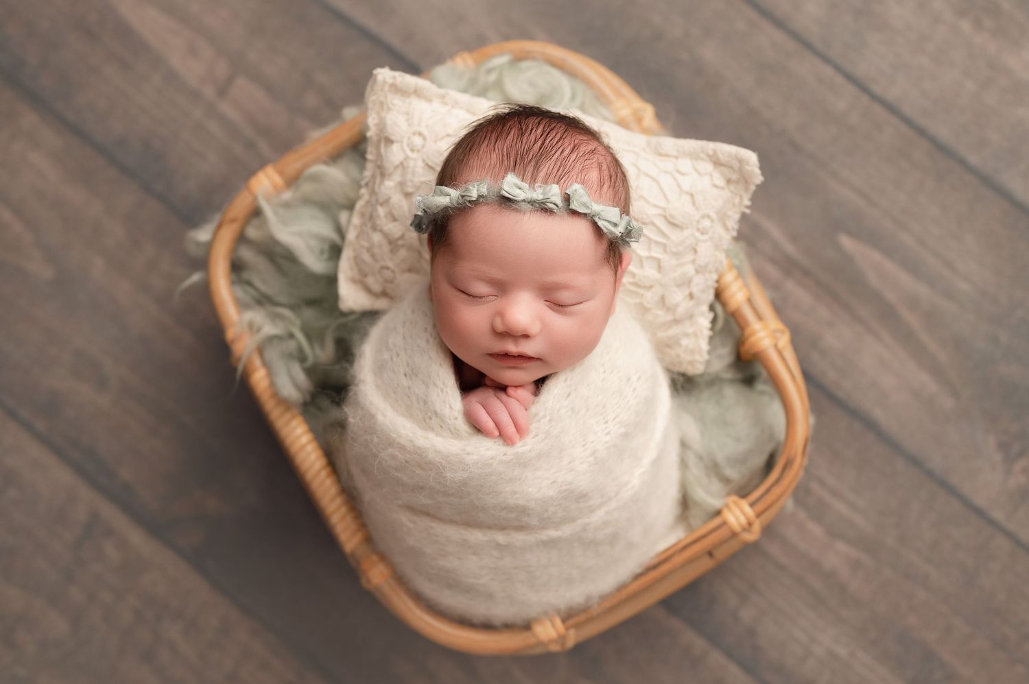 Newborn baby swaddled in cream wrap, sleeping peacefully in wicker basket with pillow and headband in my south hills photo studio.