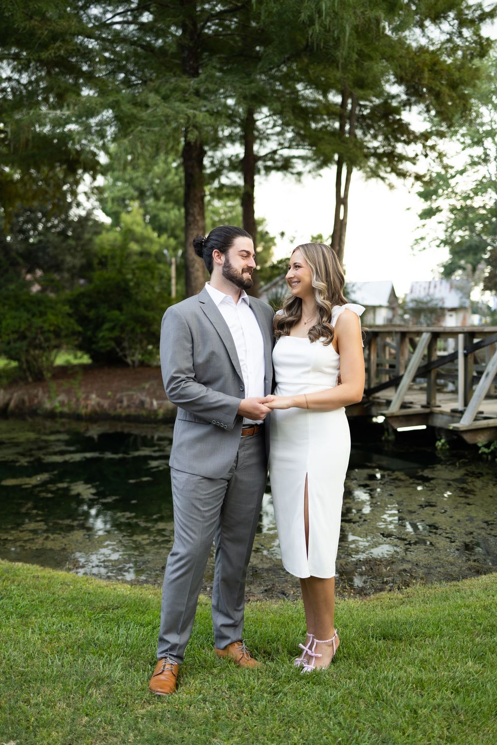 Couple in wedding attire holding hands by a pond, man in gray suit, woman in white dress.