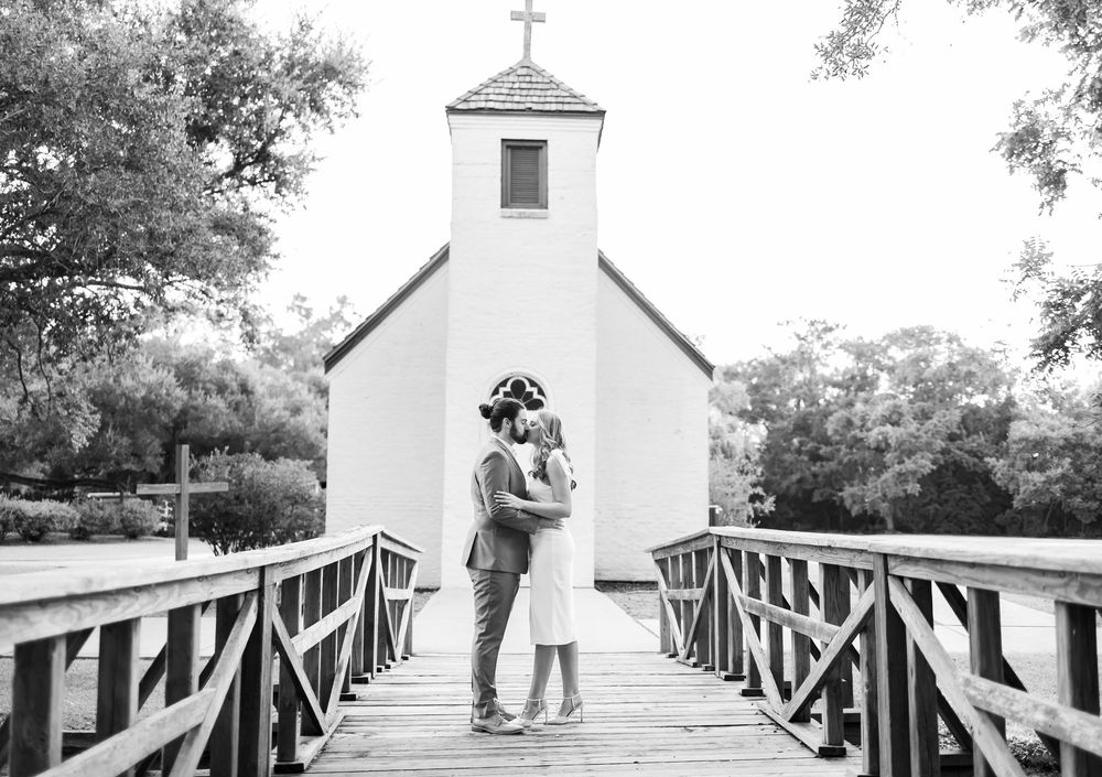 Couple kissing on wooden bridge before small white chapel in romantic black and white wedding photo.
