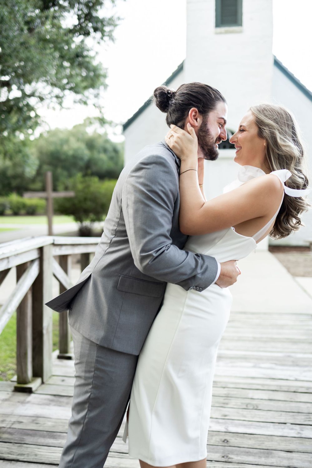 Bride and groom embracing on wooden bridge, smiling, with a white chapel in the background.