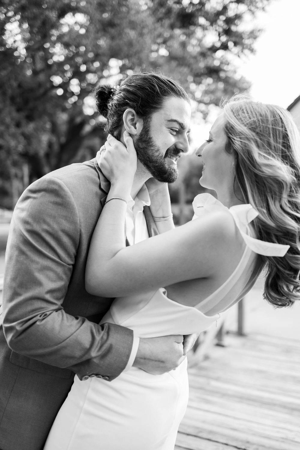 Black and white photo of a couple embracing and smiling at each other on a wooden dock, dressed in wedding attire.