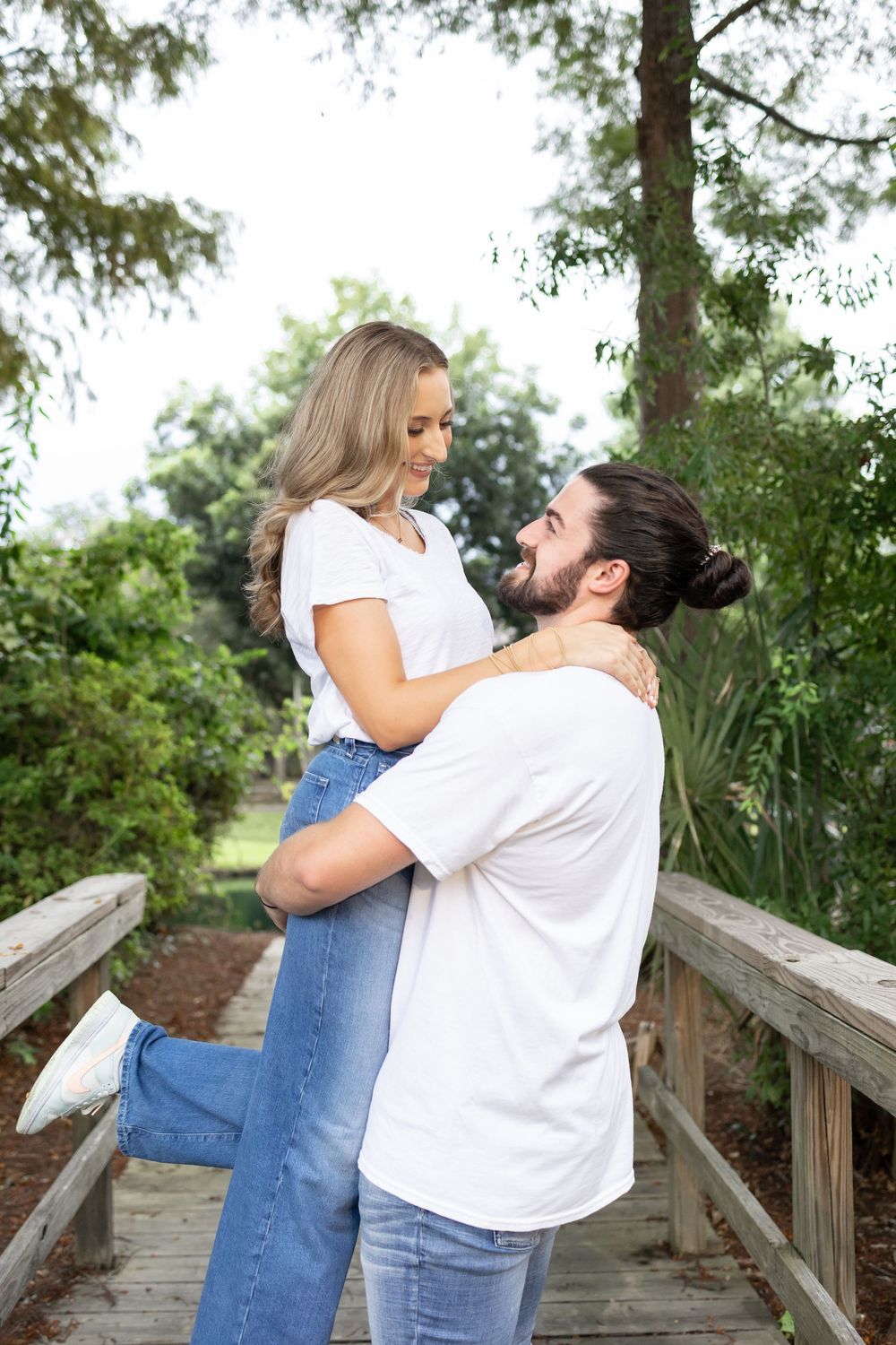 Man lifting woman on wooden bridge surrounded by lush greenery during engagement photo session.
