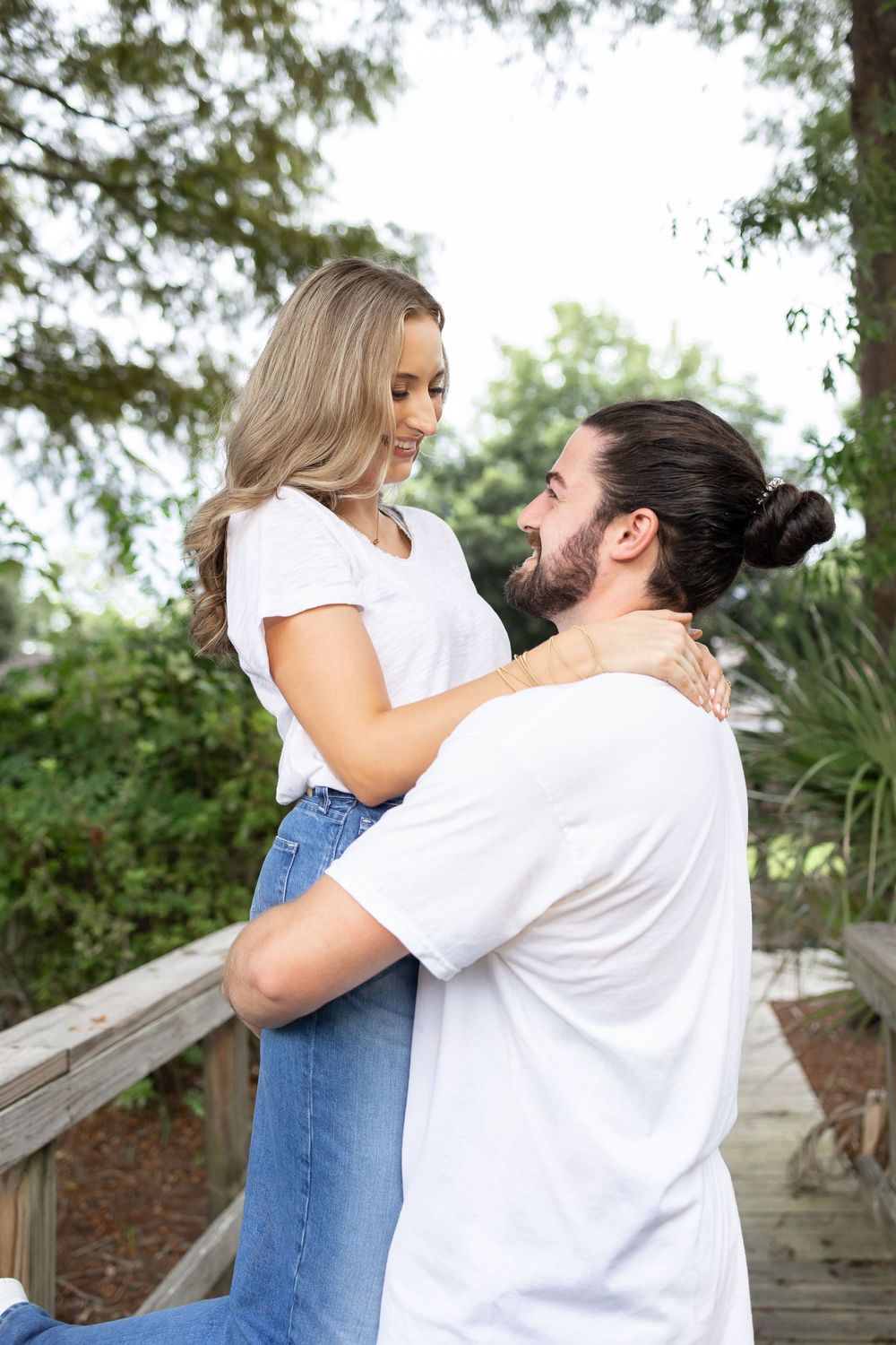 Couple embracing on wooden deck outdoors, woman in white tee and jeans, man in white shirt with bun hairstyle.
