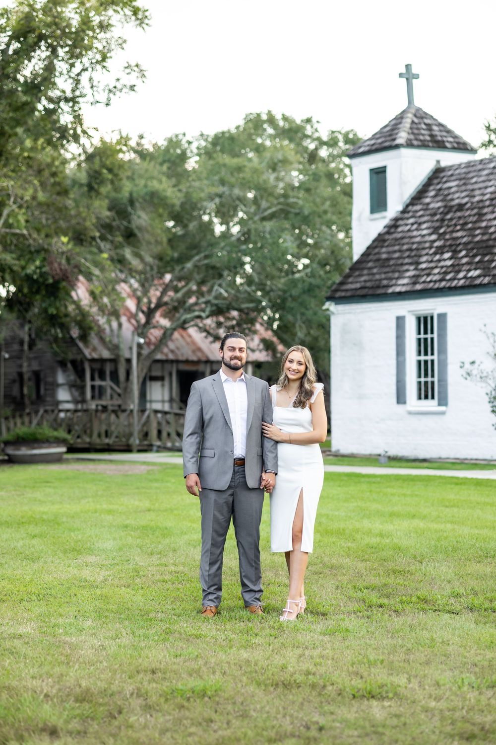 Couple posing in front of a white chapel with a cross on a lush green lawn during an outdoor engagement session.
