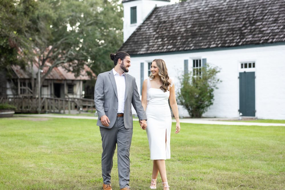 Couple walking hand in hand on green lawn before white historic building, woman in white dress, man in gray suit.