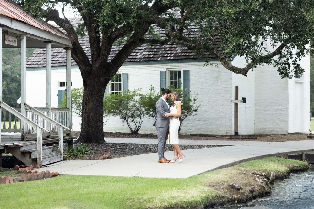 Couple embracing near white cottage building with large oak tree on lush green lawn at outdoor wedding venue.
