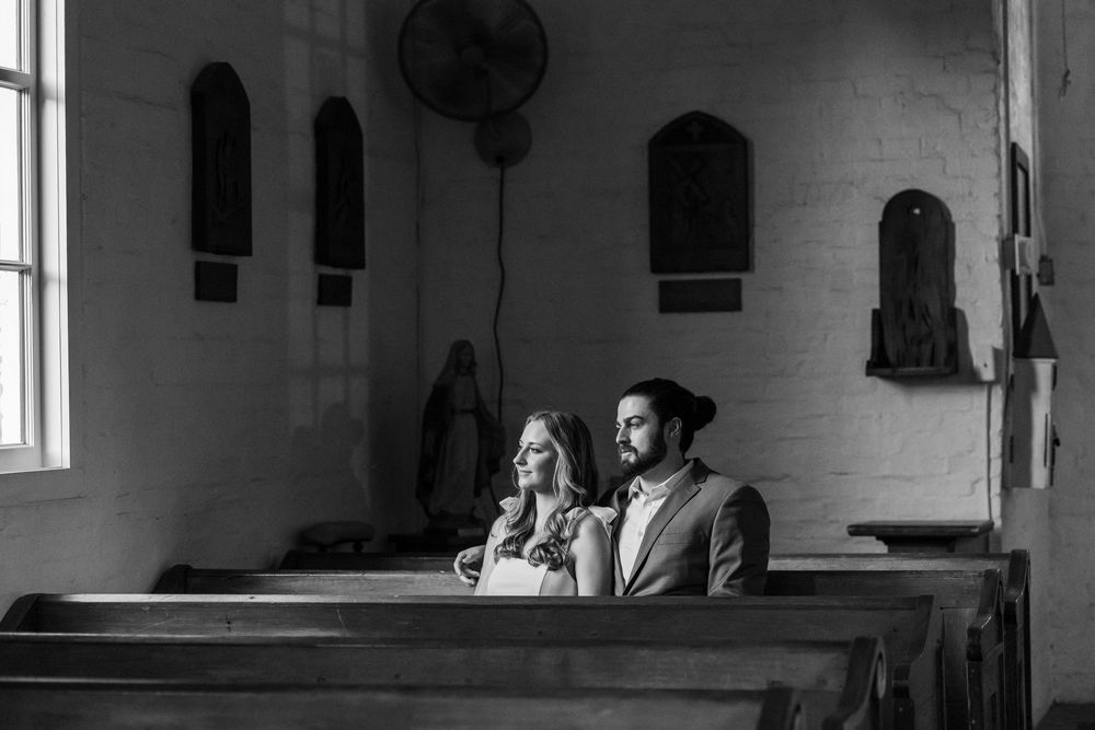 Black and white photo of a couple sitting together in a rustic church pew, bathed in soft natural light.
