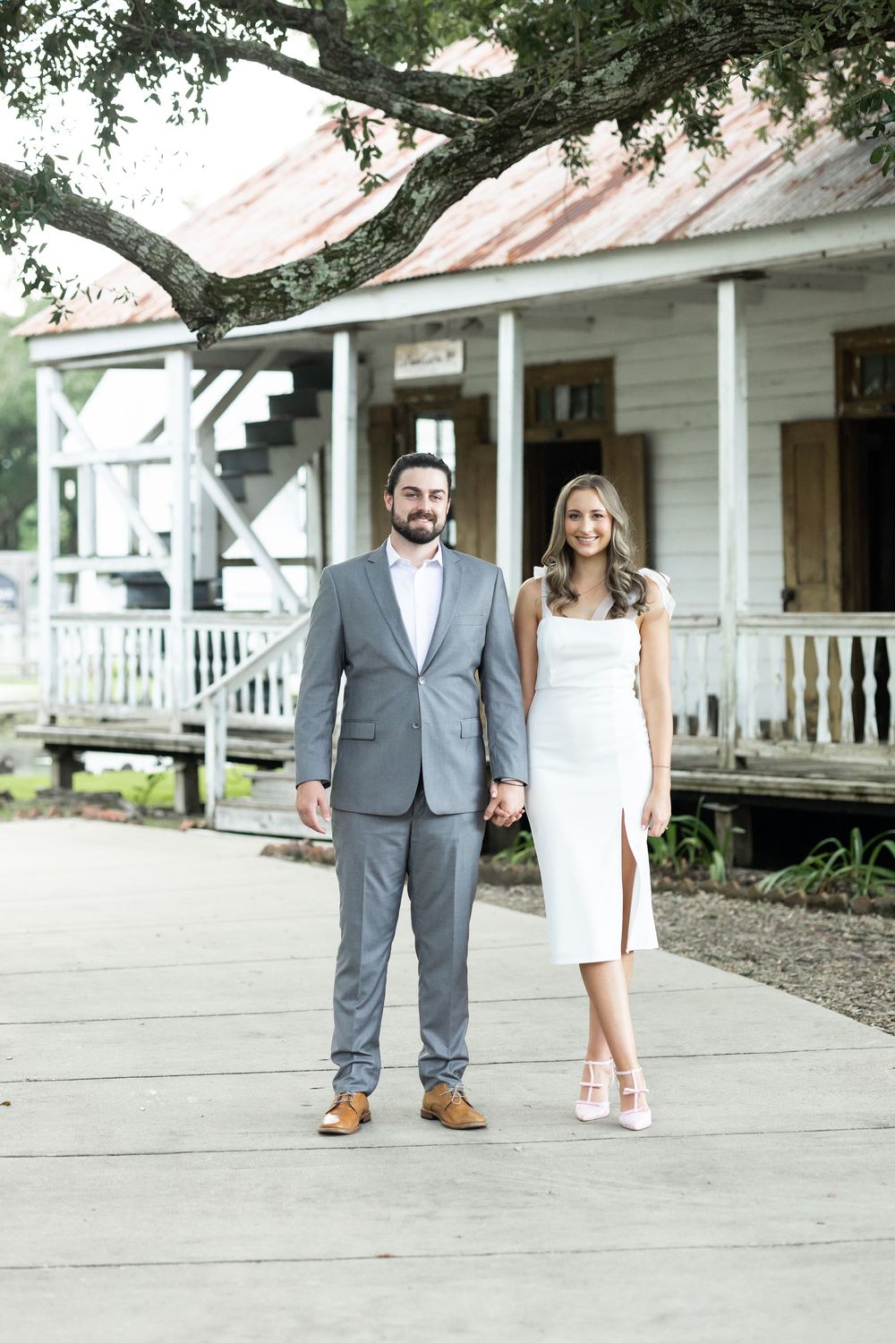 Couple posing in front of rustic white building, man in gray suit and woman in white midi dress holding hands.