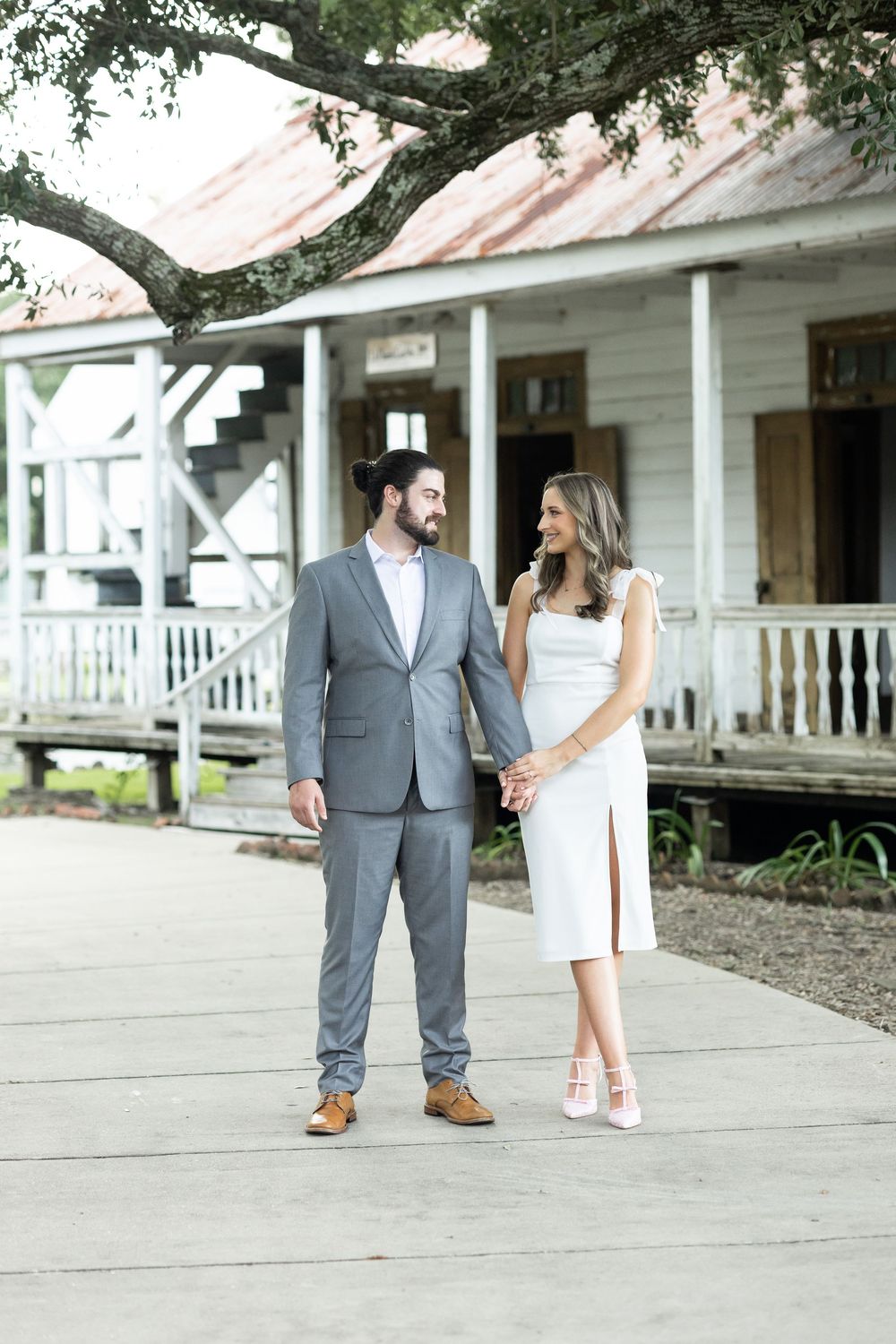 Engaged couple holding hands in white dress and gray suit outside rustic historic building.