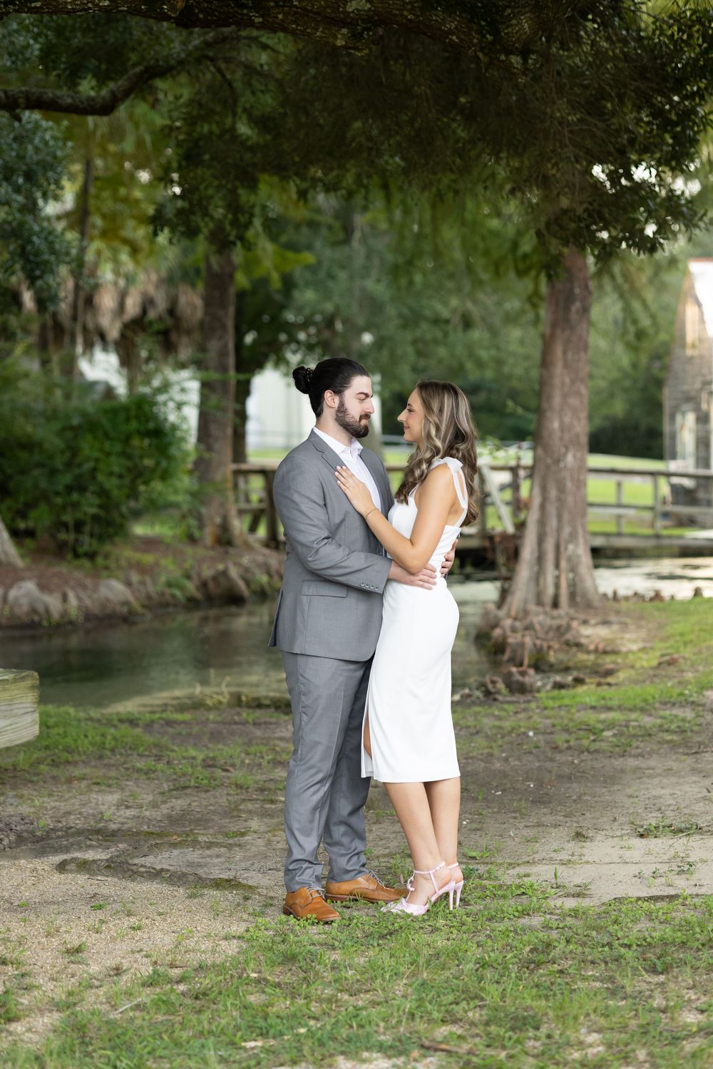 Couple in elegant attire sharing romantic moment near pond under large oak trees.