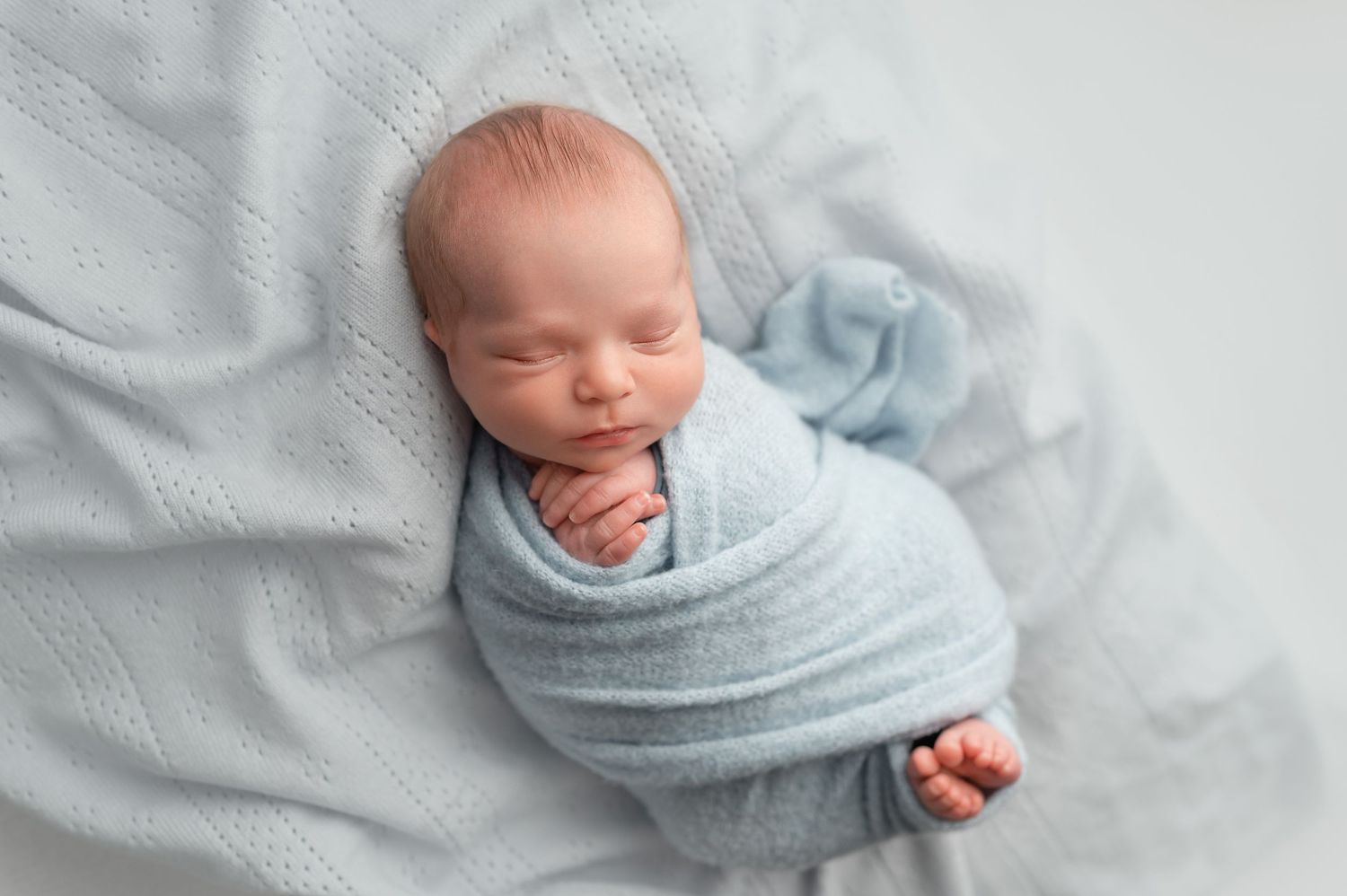 Peaceful newborn baby swaddled in a soft blue wrap, sleeping on a light gray knit blanket.