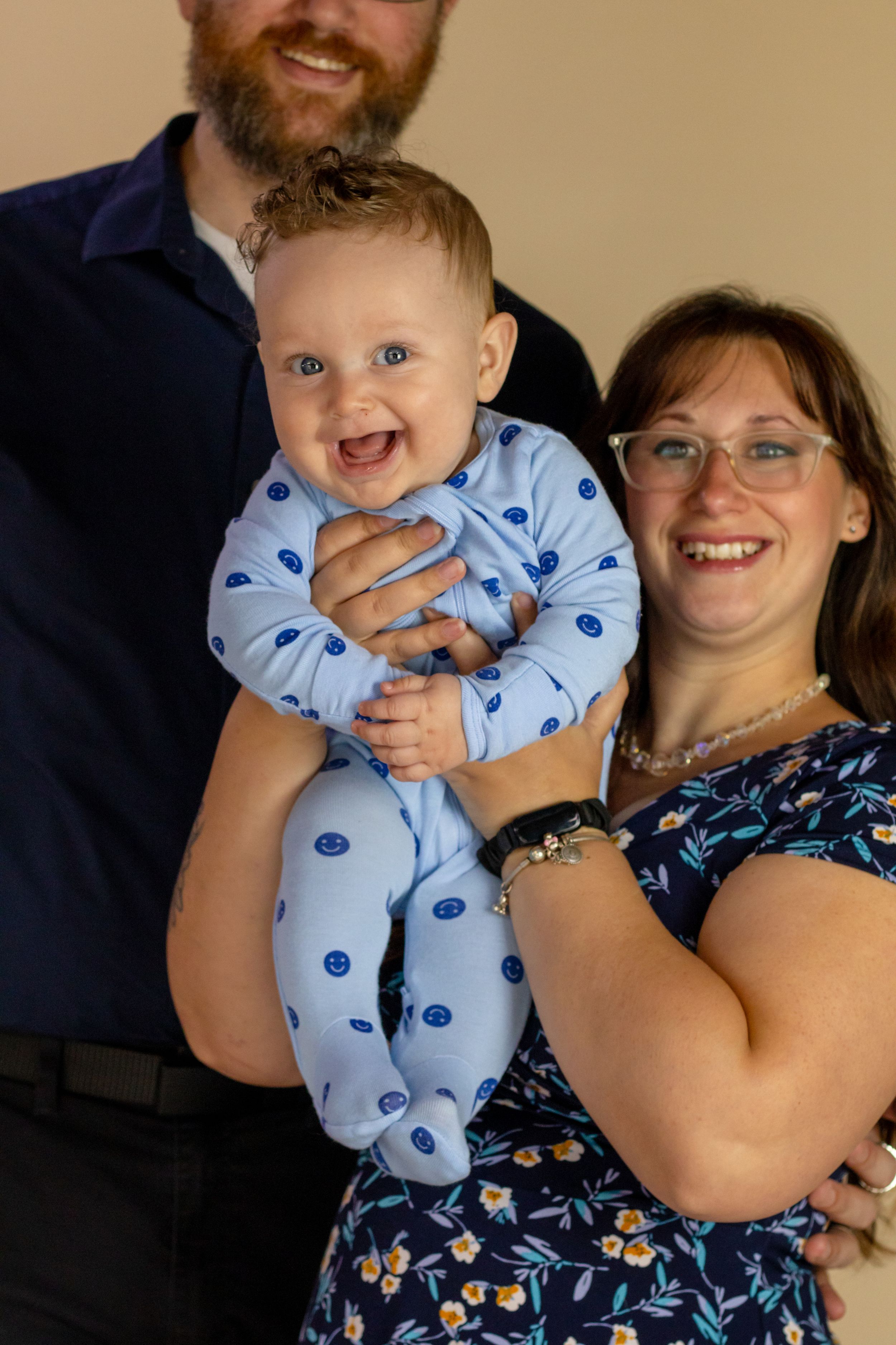 A joyful baby in a blue polka dot onesie laughs while being held by smiling parents in a studio portrait with roy washington photographer