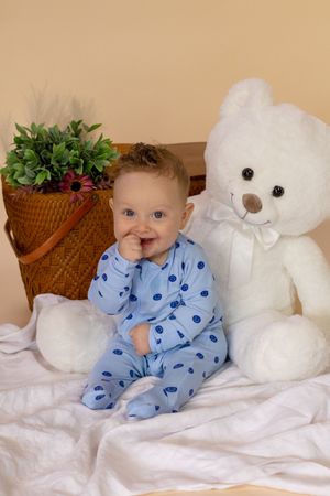An adorable baby in a blue polka dot onesie sits beside a white teddy bear next to a wicker basket with flowers in roy washington studio
