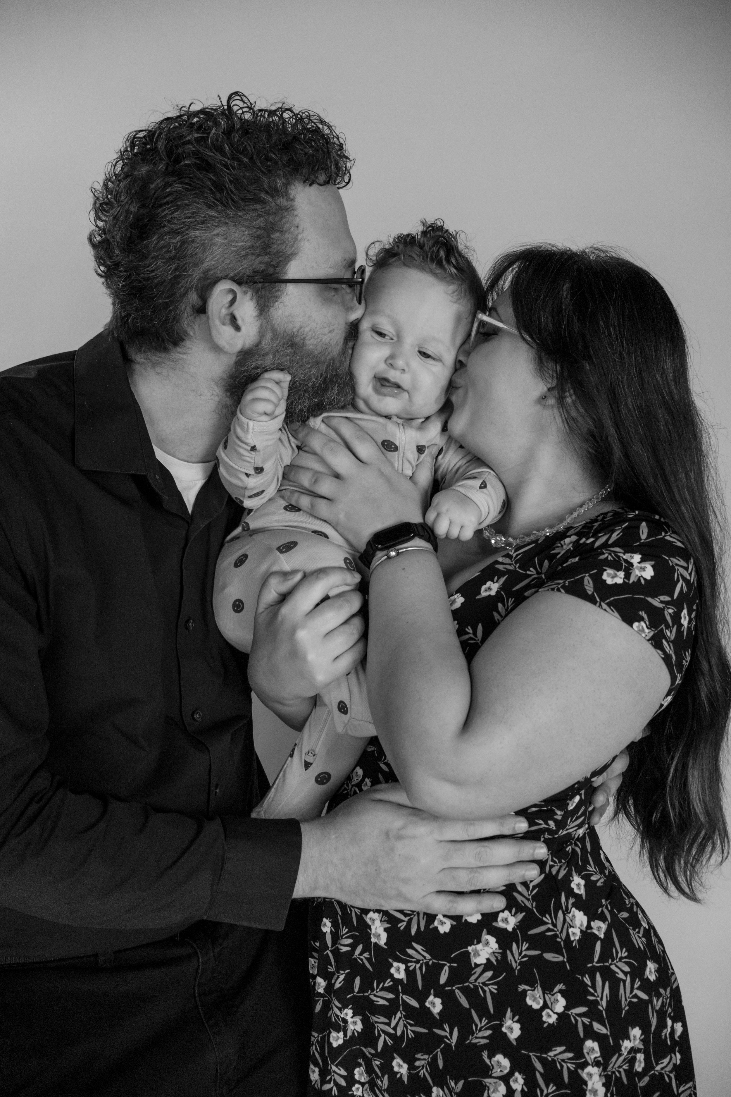 A black and white portrait of two parents lovingly kissing their baby on both cheeks taken in roy wa studio with washington photographer