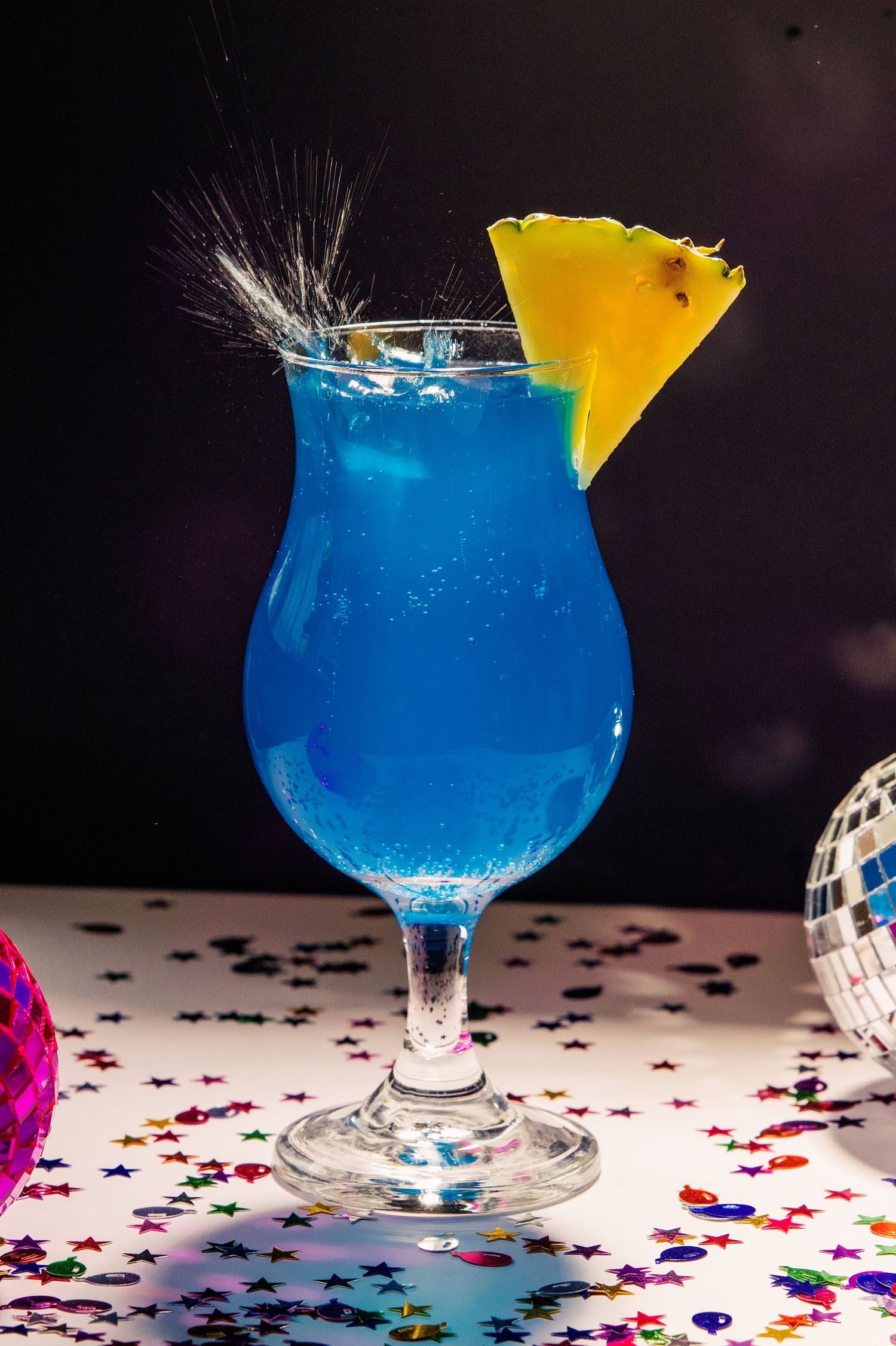 Blue lagoon cocktail with pineapple garnish in hurricane glass, surrounded by star confetti and disco balls.