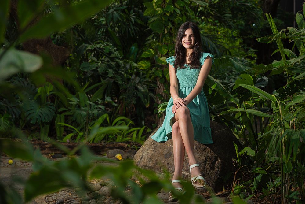 Young man in white t-shirt and shorts sitting on a log surrounded by lush tropical green foliage.