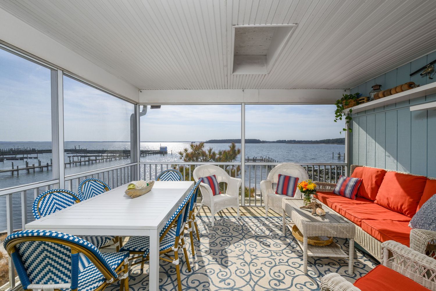 Screened porch with ocean views, white dining table, blue chairs, orange sofa, and patterned rug overlooking a waterfront dock.