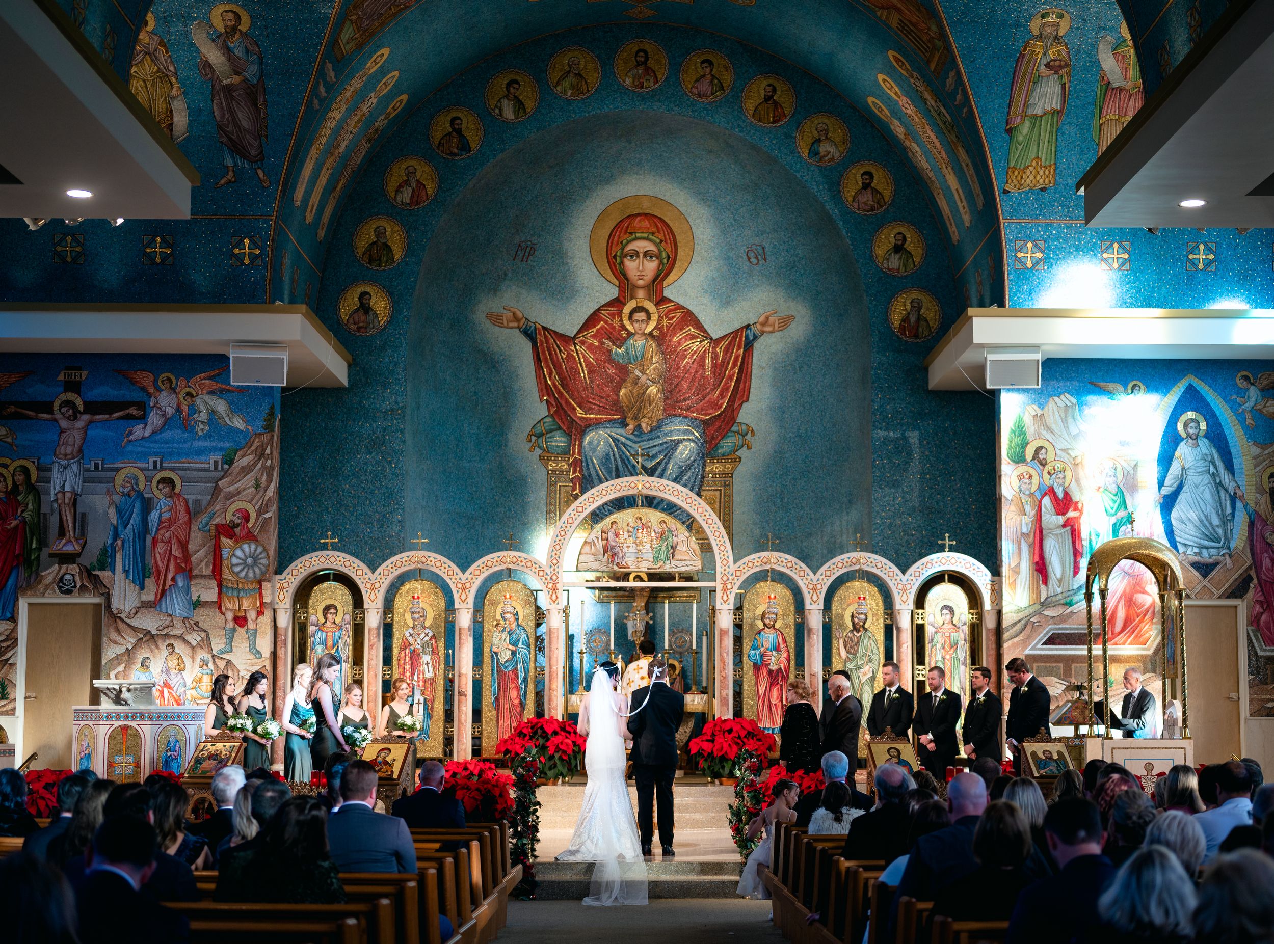 Wedding ceremony in ornate Byzantine Orthodox church with colorful religious murals and icons adorning the walls and altar.