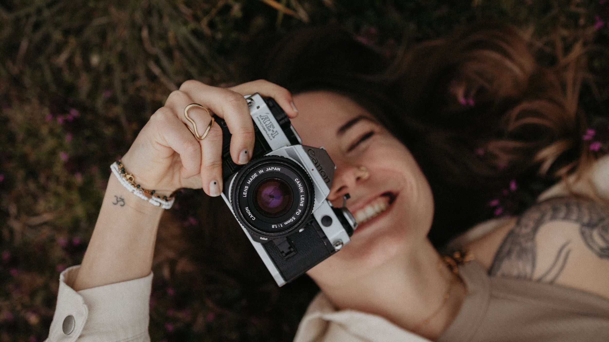 Smiling woman lying on grass holding a vintage film camera, with tattoos and jewelry visible.