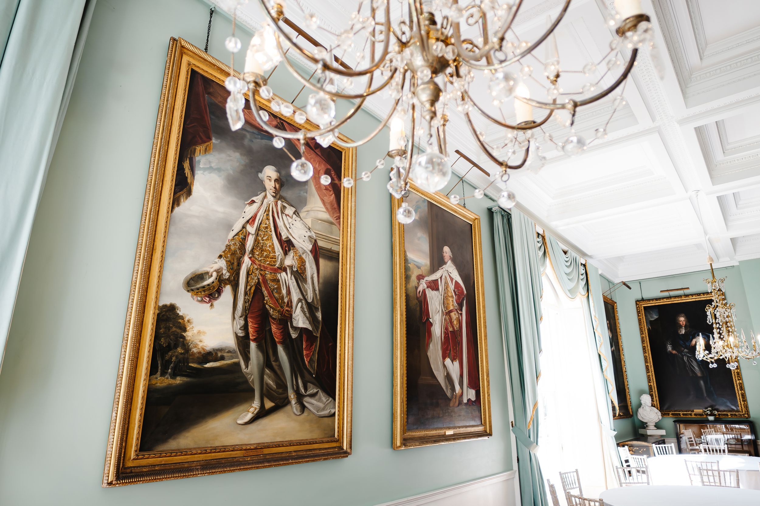 Elegant room with gold-framed portrait paintings of aristocrats on mint green walls beneath an ornate crystal chandelier.