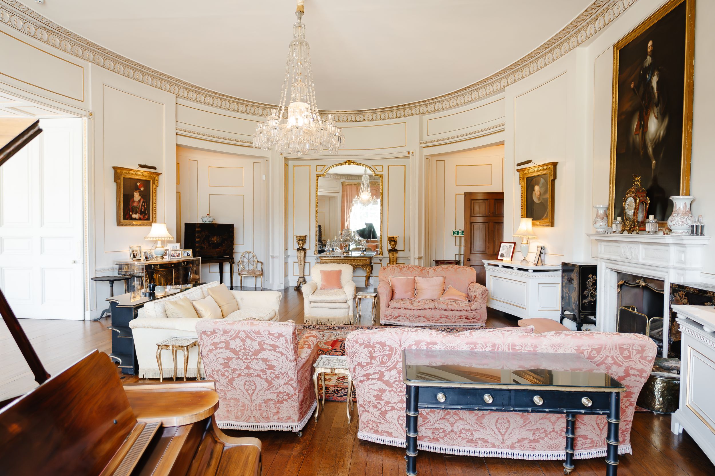 Elegant oval drawing room with pink upholstered sofas, crystal chandelier, ornate fireplace, and hardwood floors in a grand estate.