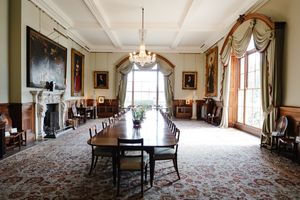 Elegant formal dining room with long mahogany table, chandelier, ornate fireplace, portrait paintings, and draped windows.