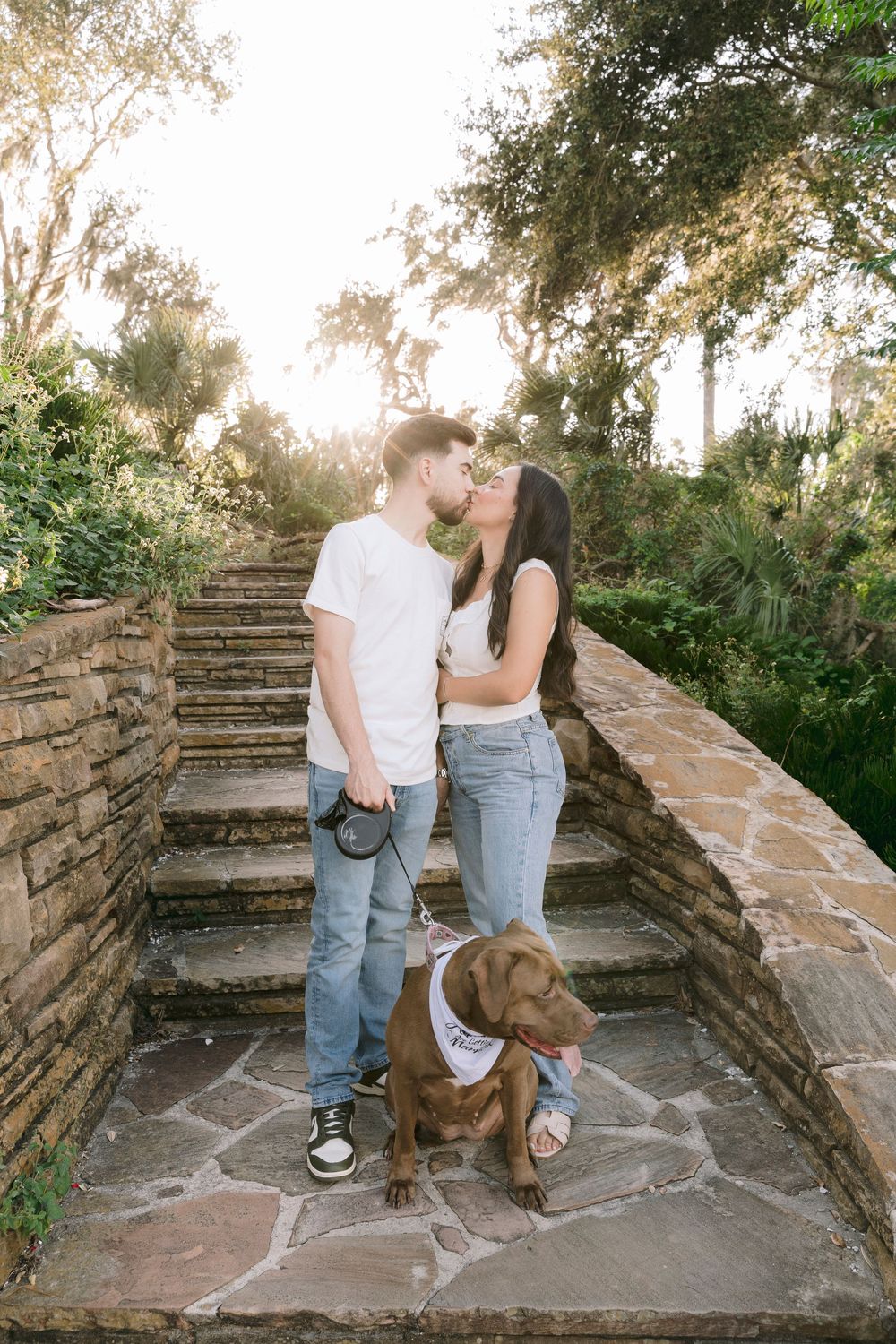 Couple sharing a kiss on stone steps with their brown Labrador wearing a bandana, surrounded by lush greenery at sunset.
