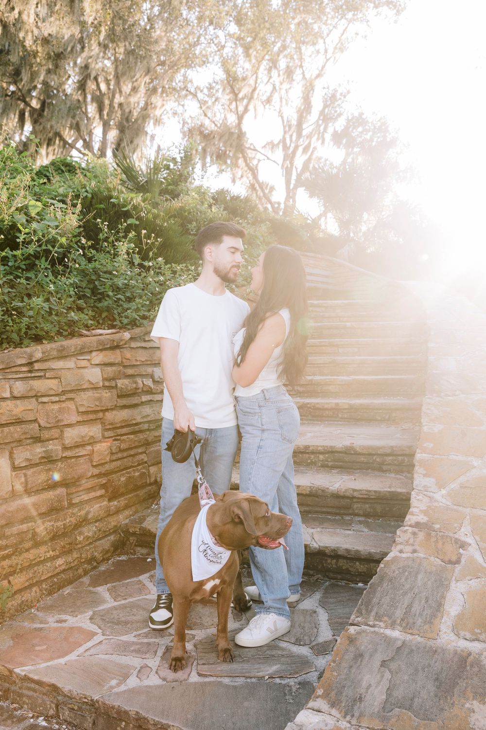 Couple sharing a romantic moment with their brown dog on stone steps in a sun-flared garden.