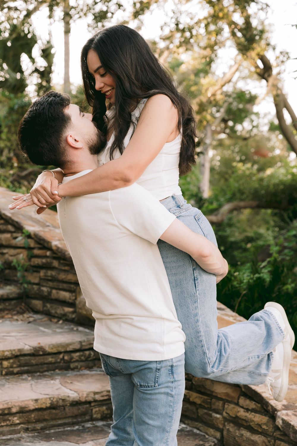 Man lifting woman in his arms outdoors on stone steps surrounded by lush greenery.