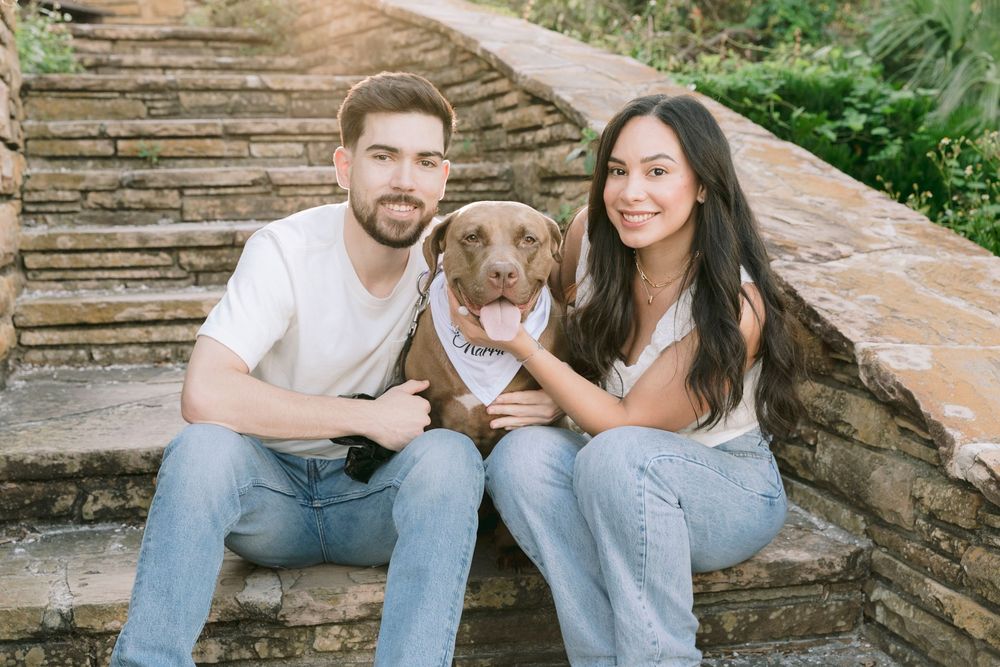 Couple sitting on stone steps with a brown dog wearing a bandana, surrounded by lush greenery.
