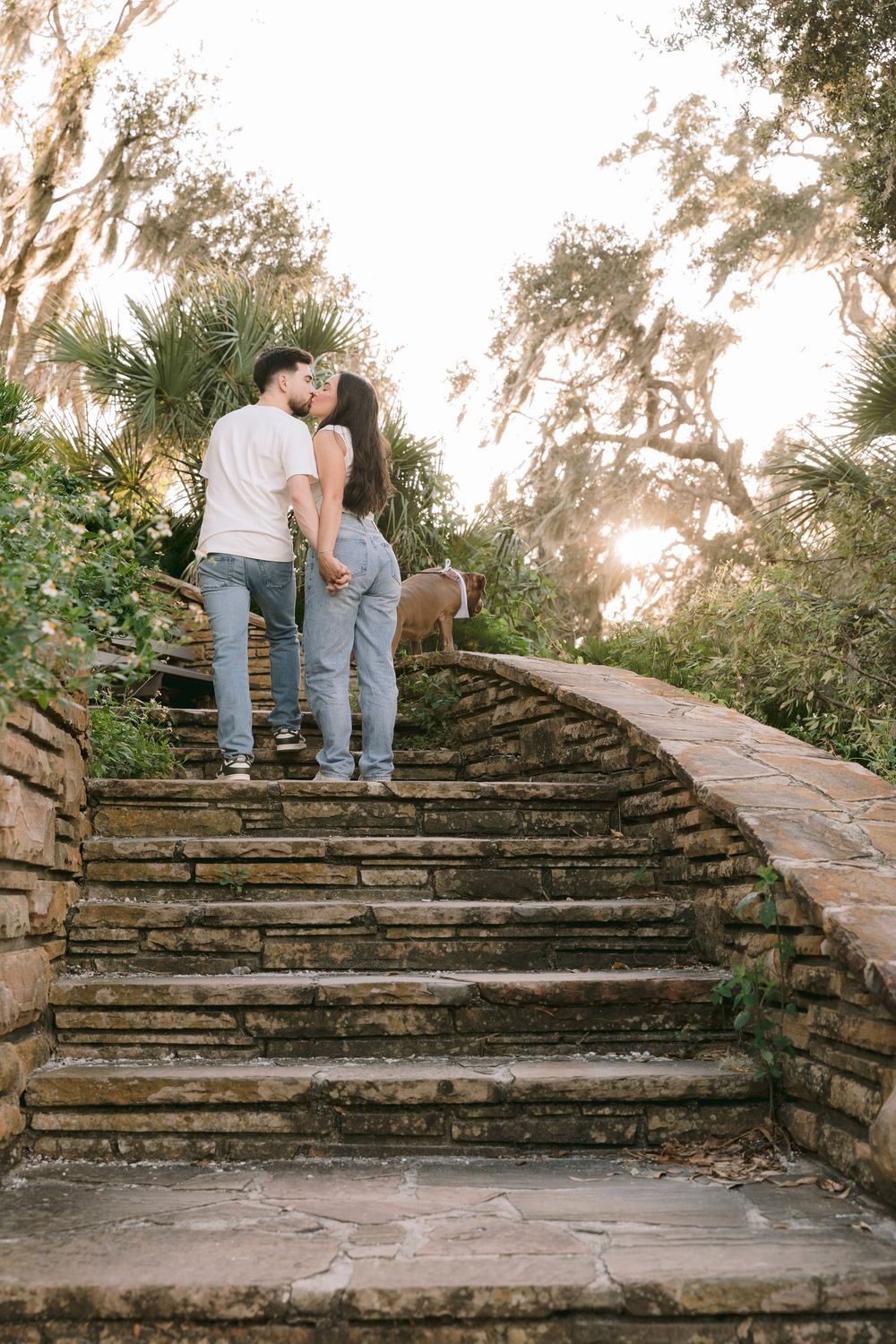 Couple kissing on stone steps at sunset with their dog in a scenic garden setting.