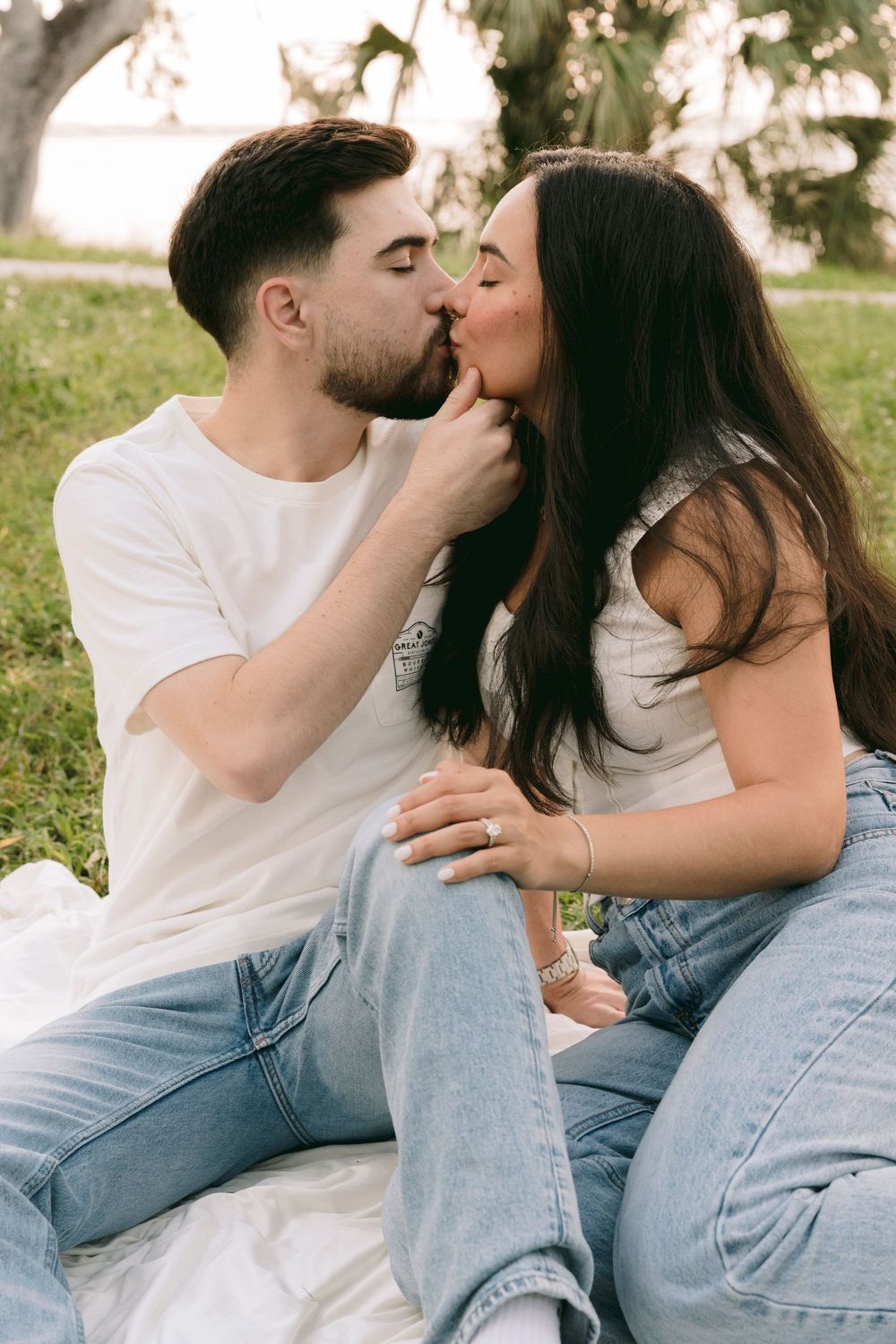 A couple sharing a romantic kiss while sitting on a blanket in a park wearing matching white tops and blue jeans.