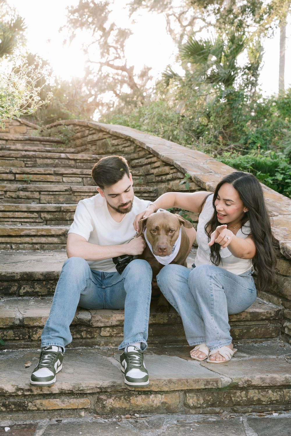 Couple sitting on stone steps with their brown dog in a sunlit outdoor park setting.
