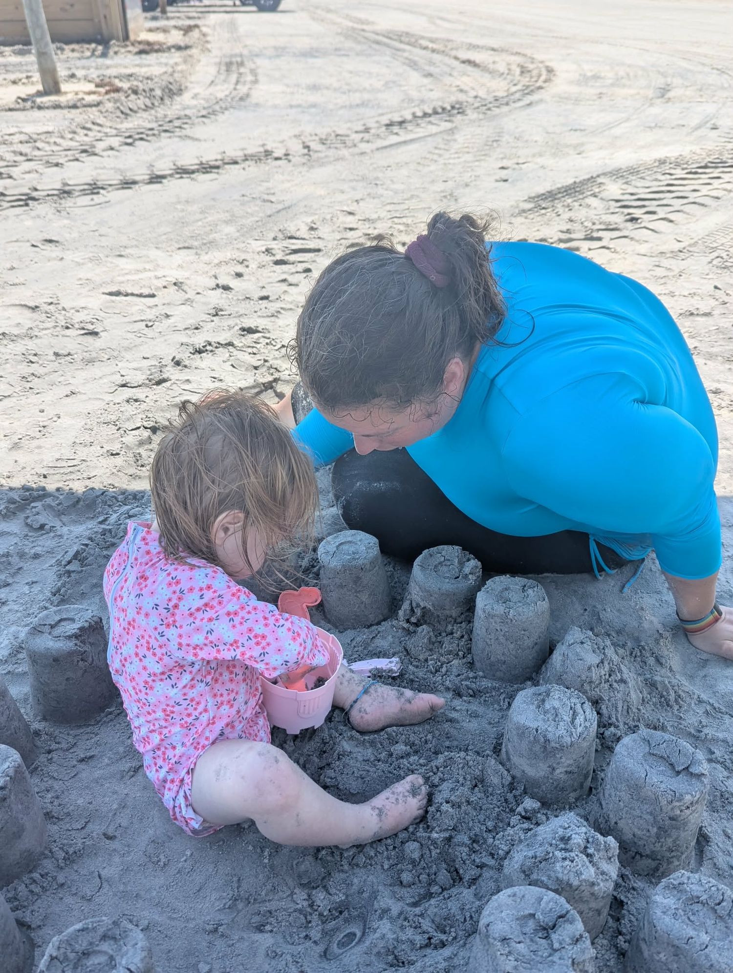 Woman in blue shirt and toddler girl building sand castles together on a sandy beach.
