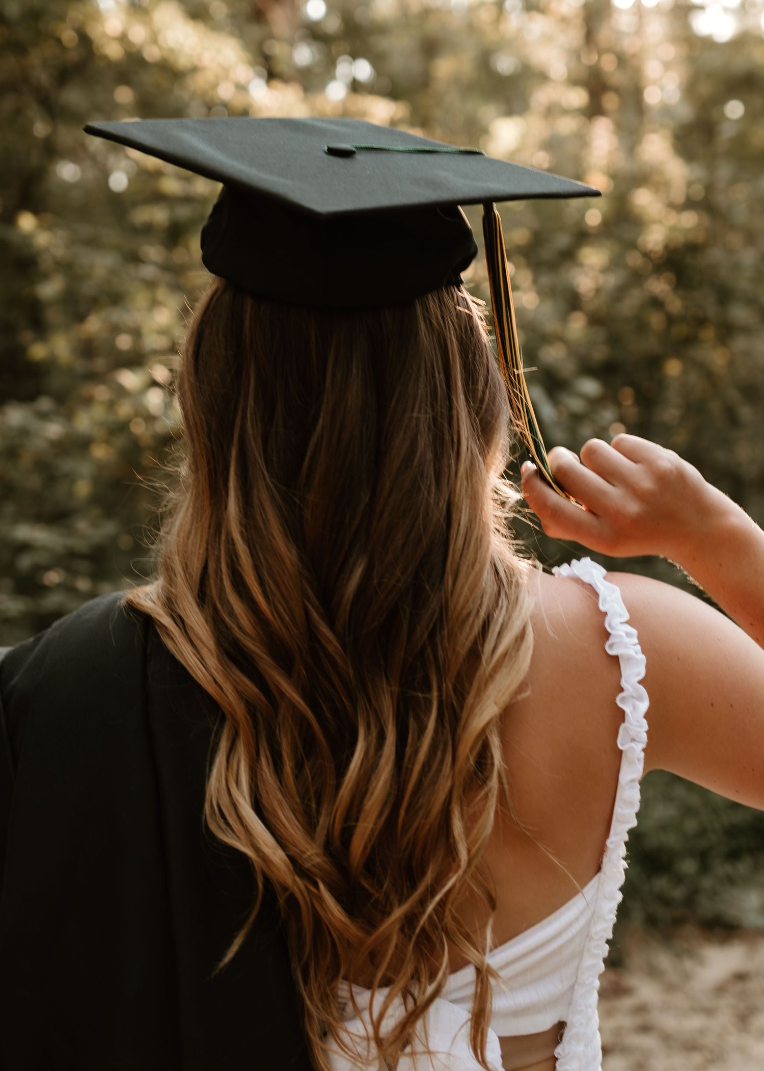 Graduate in black cap and gown with long wavy hair poses outdoors among lush green trees.
