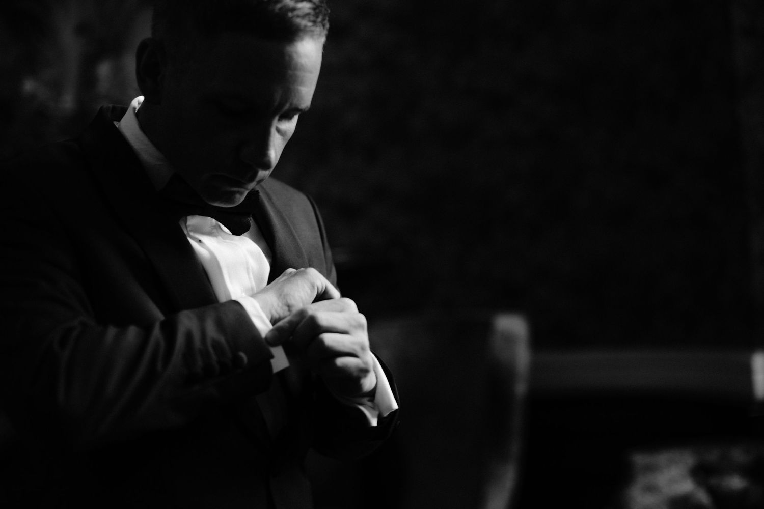 Black and white photo of a man in a tuxedo adjusting his cufflinks, elegant wedding preparation moment.