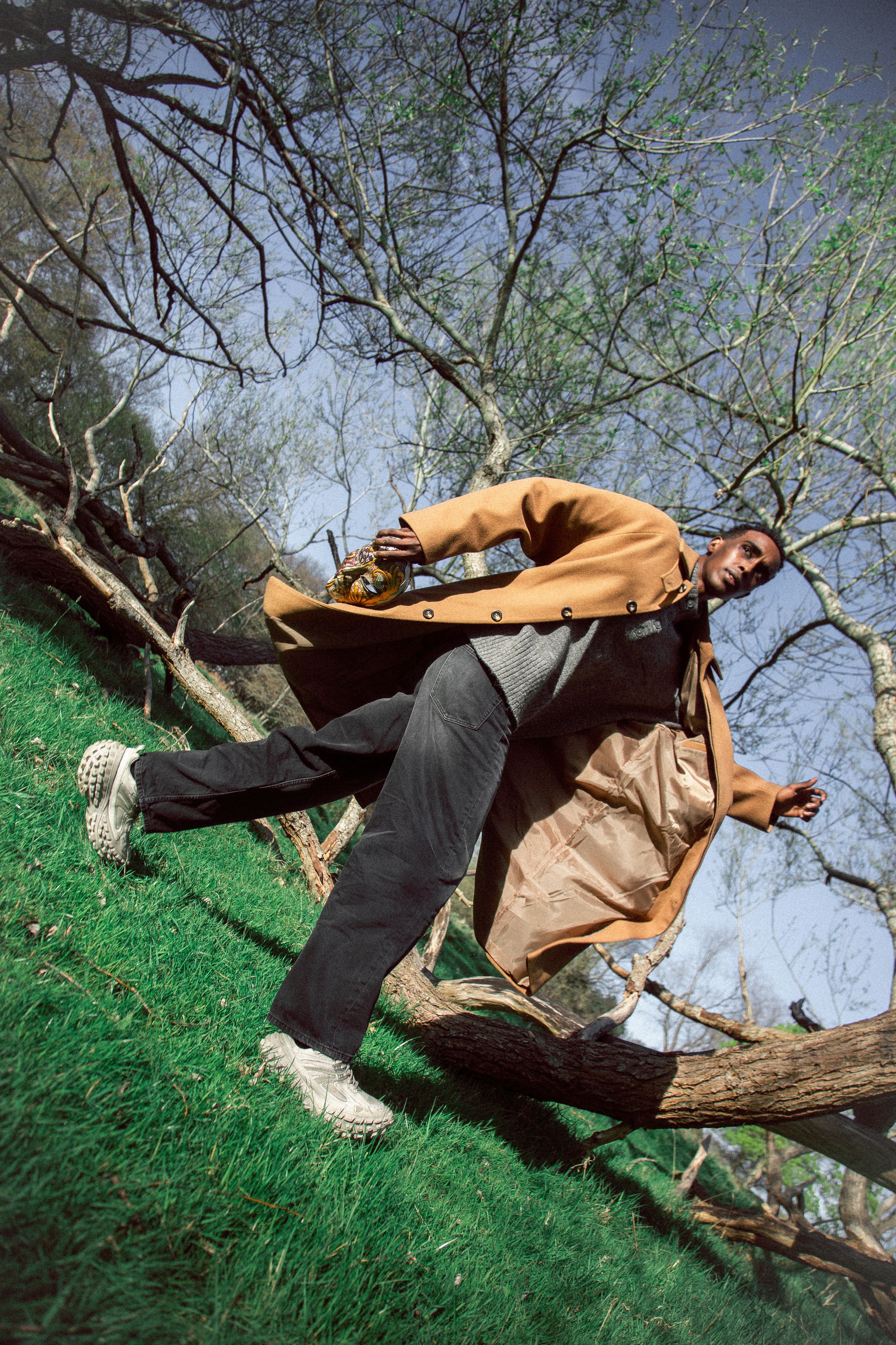 Man in orange coat leaning on fallen tree branch on grassy hill with bare trees in background.