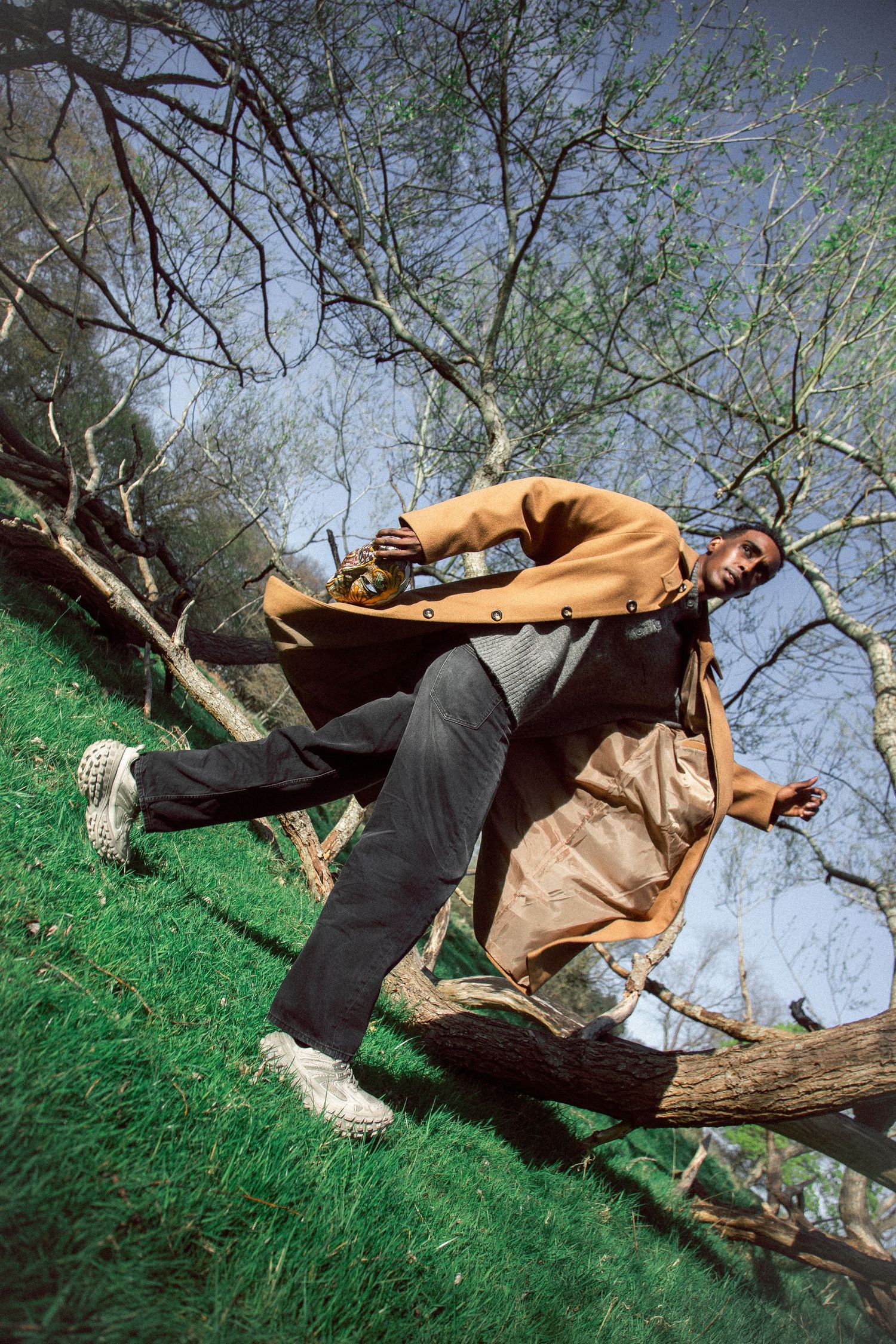 Man in orange coat leaning on fallen tree branch on grassy hill with bare trees in background.
