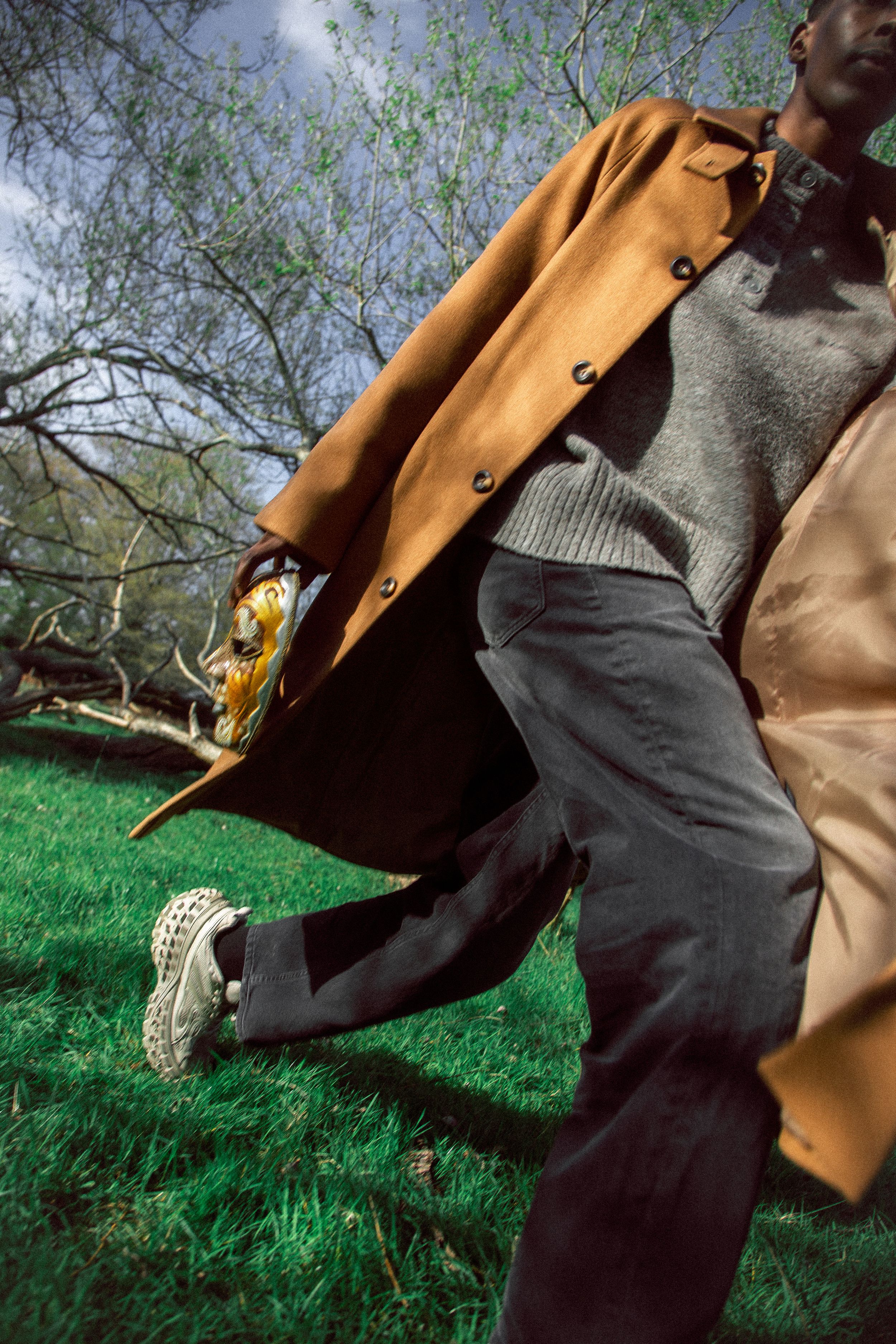 Man in camel coat, grey sweater, dark trousers, and white sneakers walking on green grass outdoors.