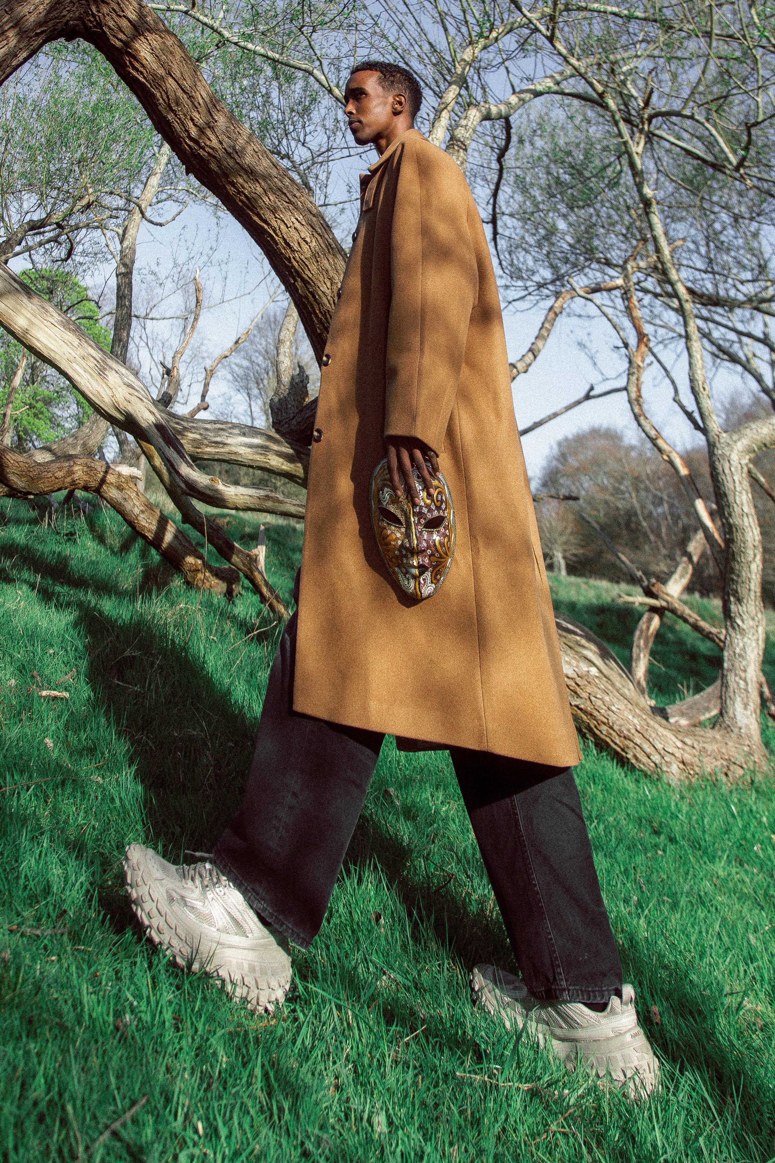 Man in long camel coat holding decorative mask, wearing dark trousers and white sneakers in a park.