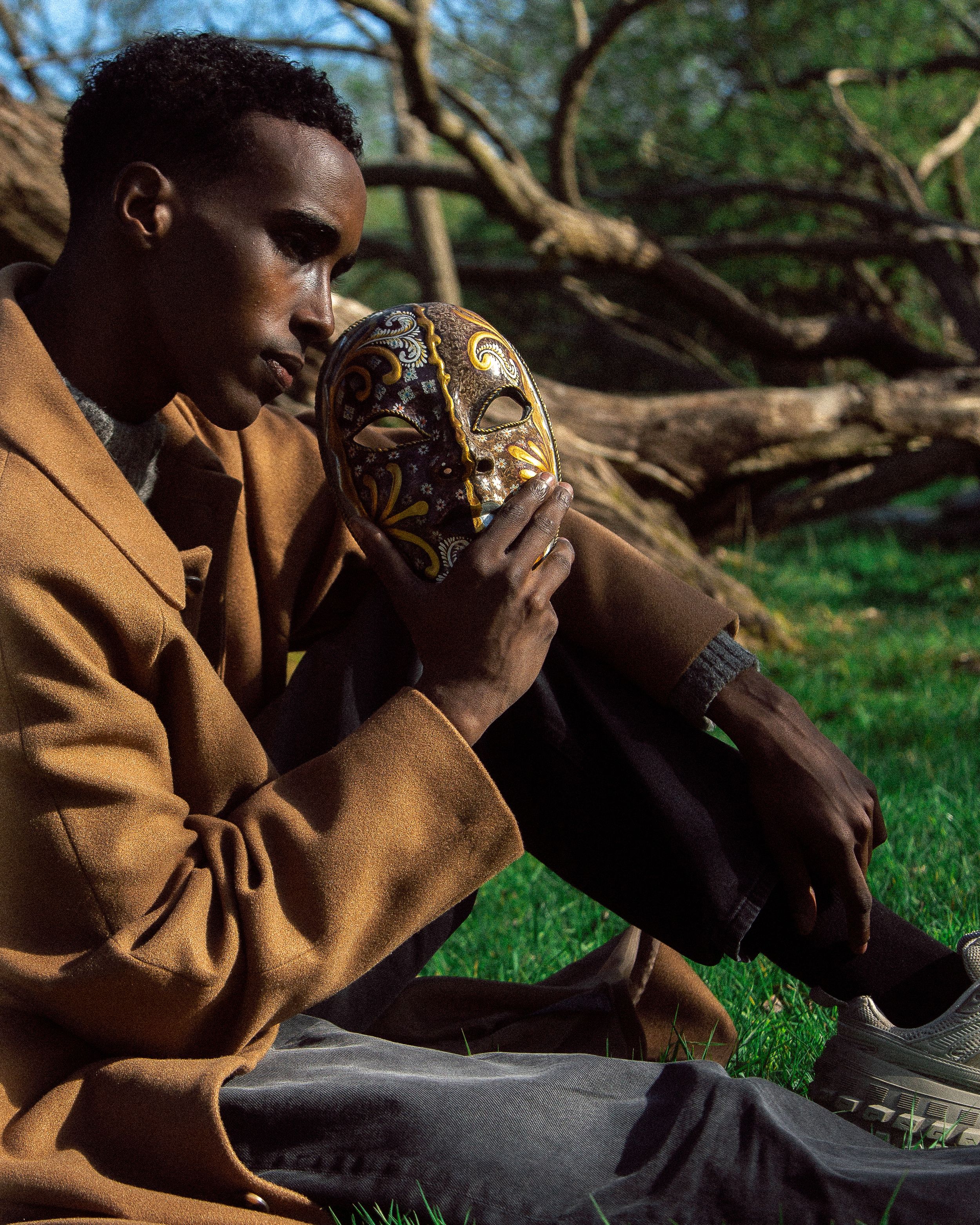 Man in tan blazer holding ornate decorative mask while sitting outdoors near fallen trees on green grass.