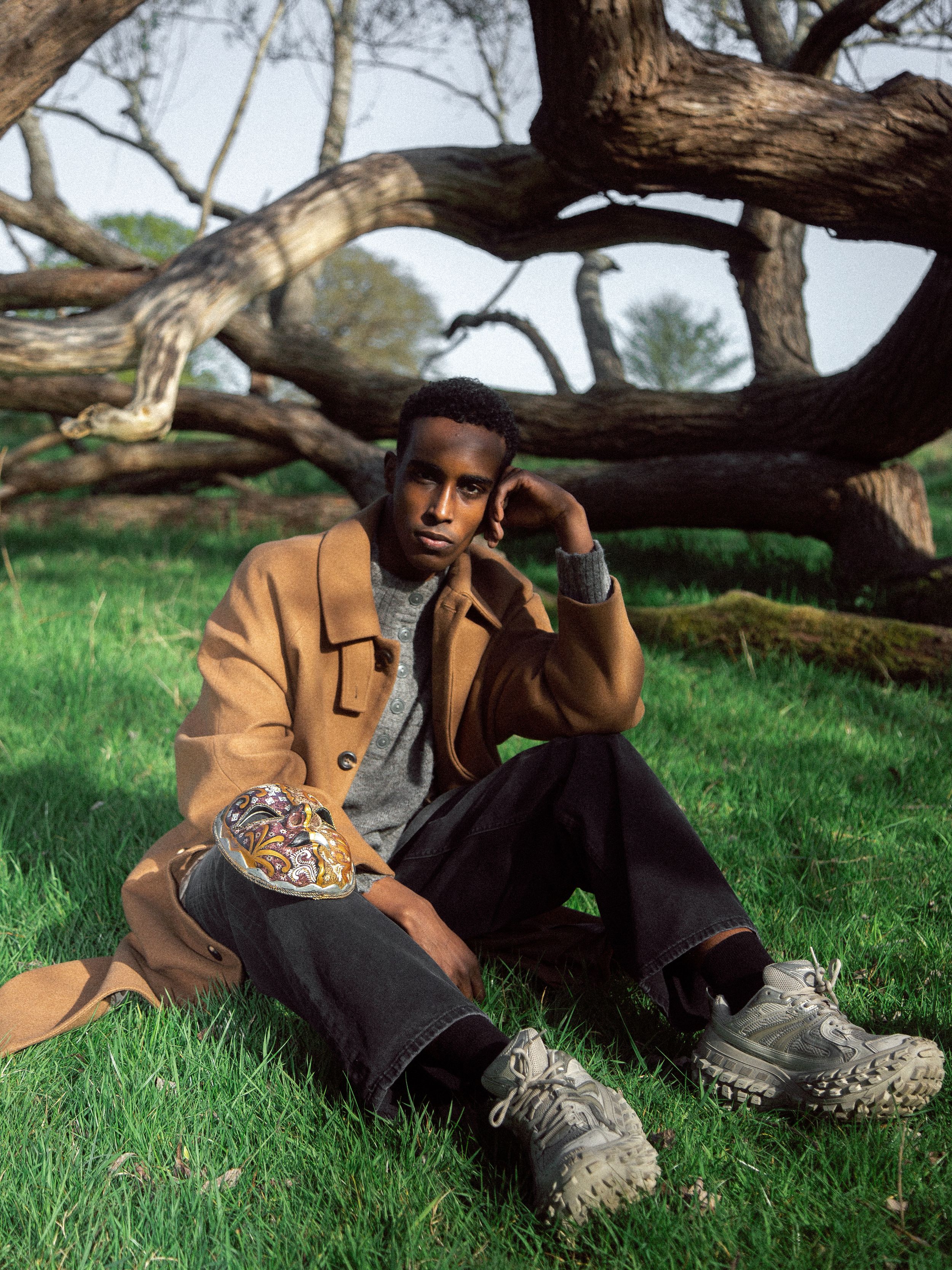 Young man in camel coat and dark trousers sits on grass leaning against a large tree in a natural outdoor setting.