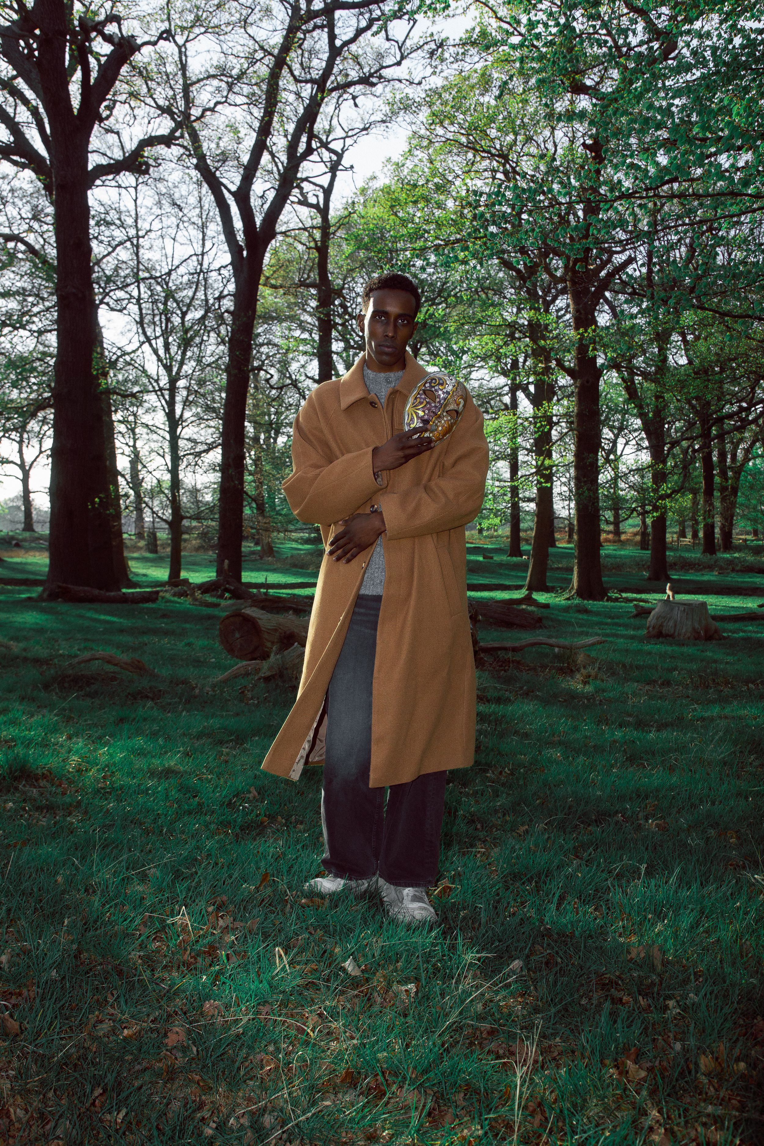 Man in camel trench coat holding flowers stands in a lush green forest clearing with tall trees.