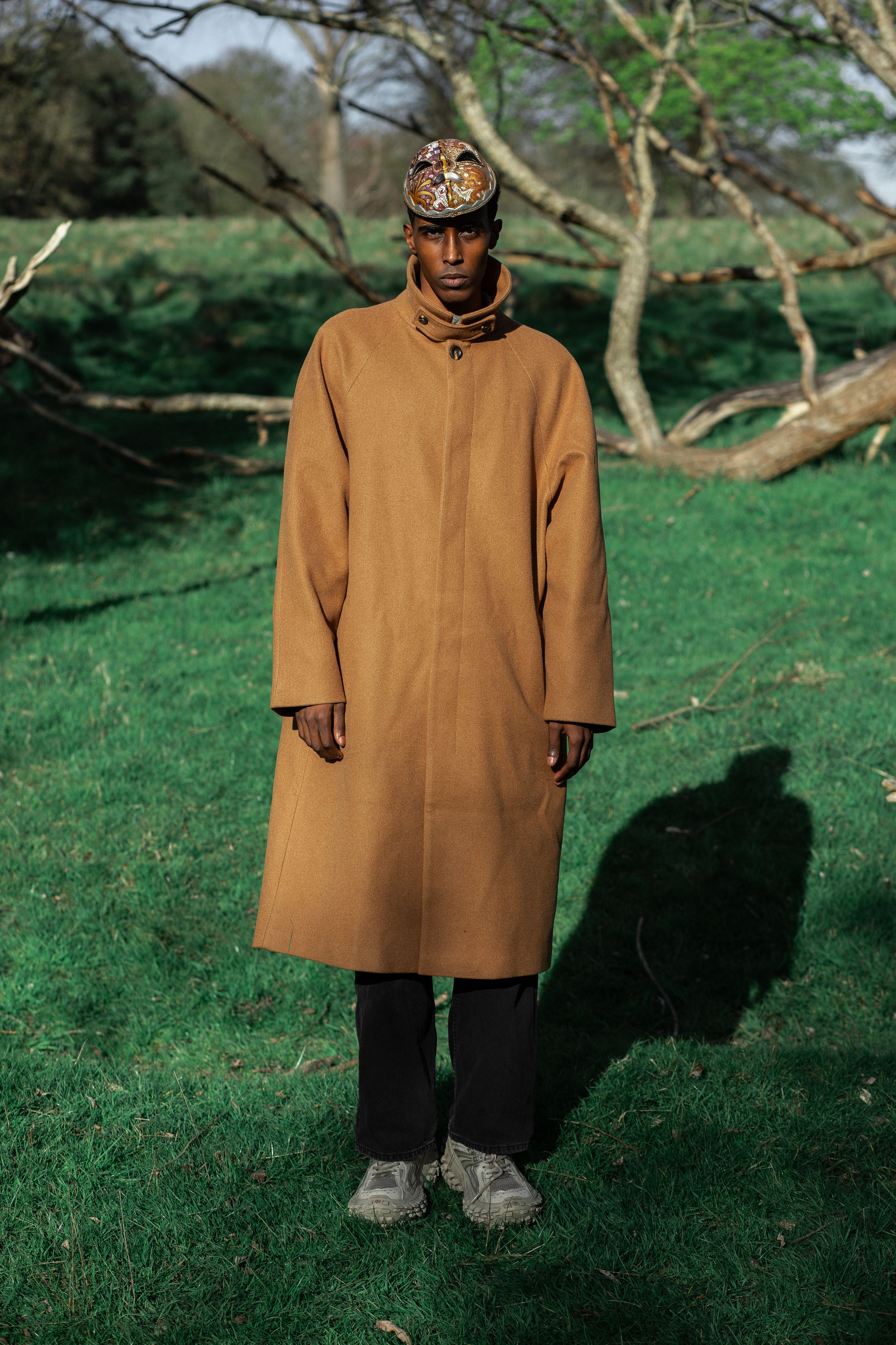 Man in camel coat and embroidered cap standing on green grass with bare trees in background.