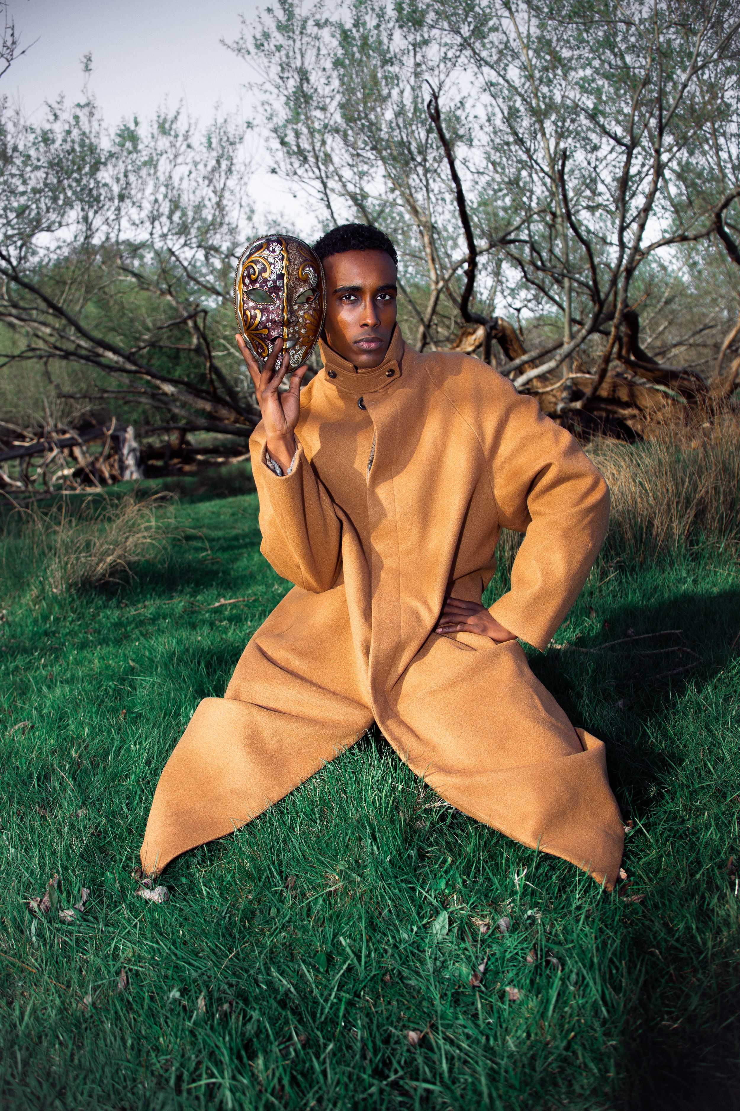 Man in tan outfit poses on grass holding decorative mask near trees.